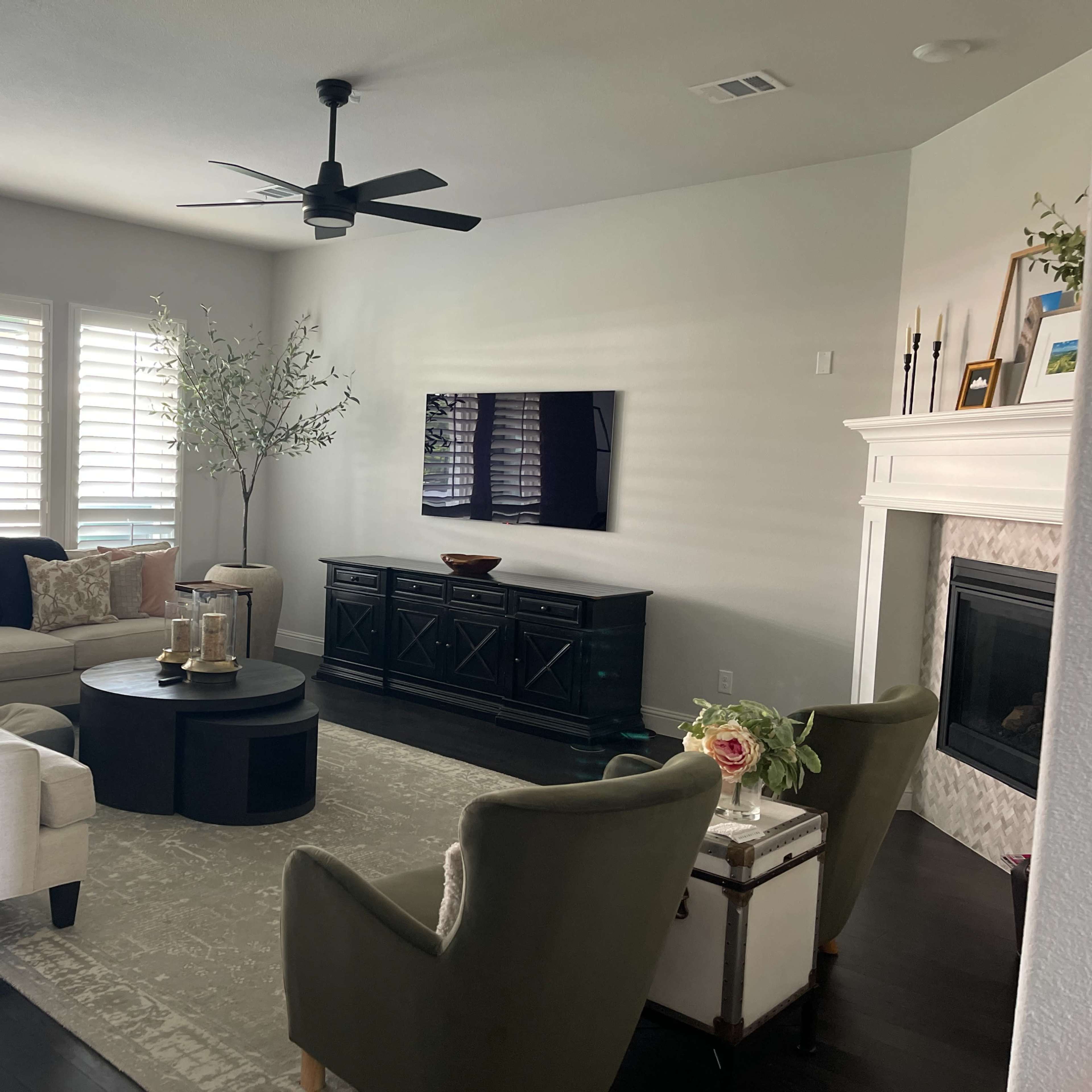 The living room features a light-colored sectional sofa, a round coffee table, a television mounted on the wall, and a mix of decorative elements, such as a plant and flowers.