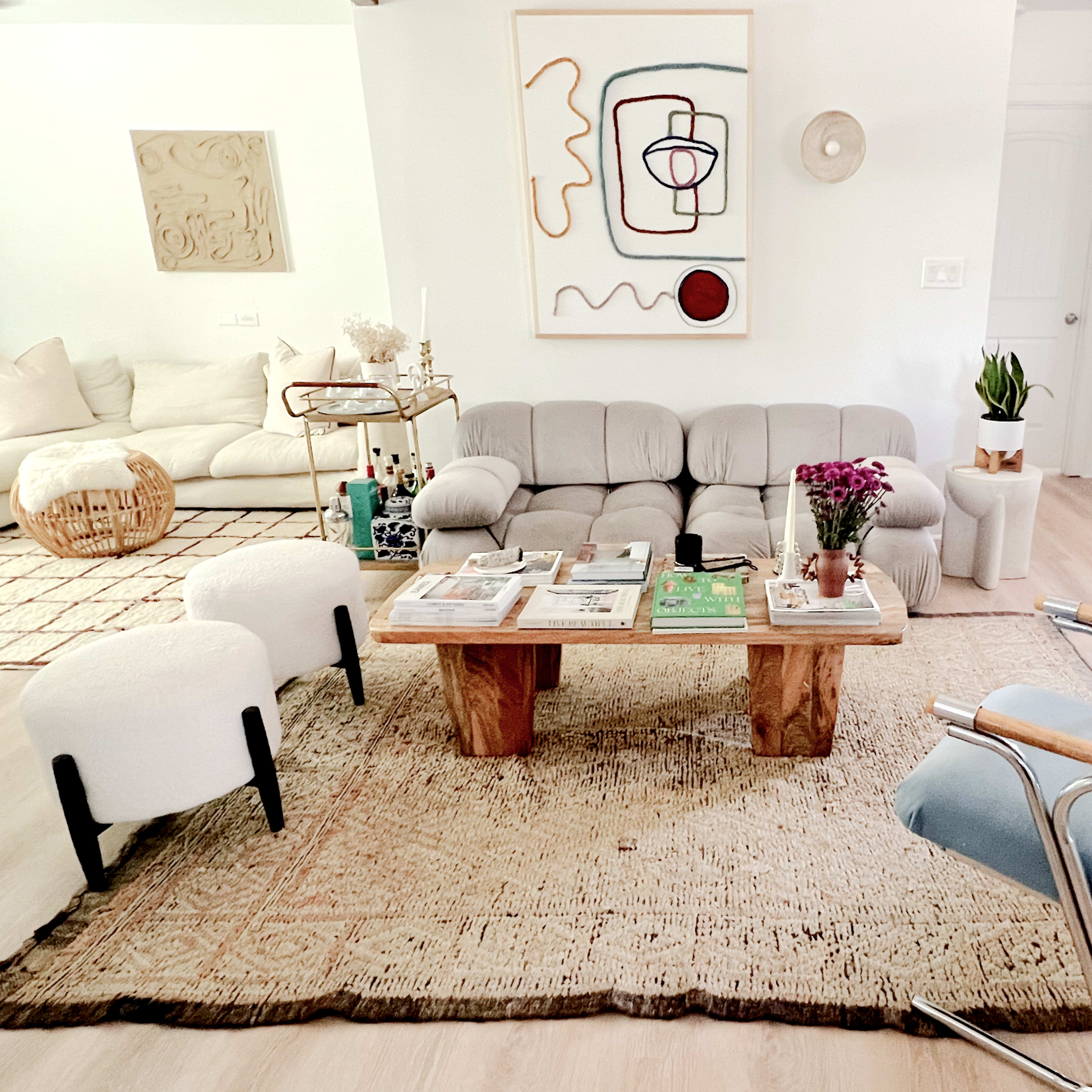 A modern living room featuring a light-colored sofa, a wooden coffee table with magazines, two minimalist armchairs, and decorative art on the walls.