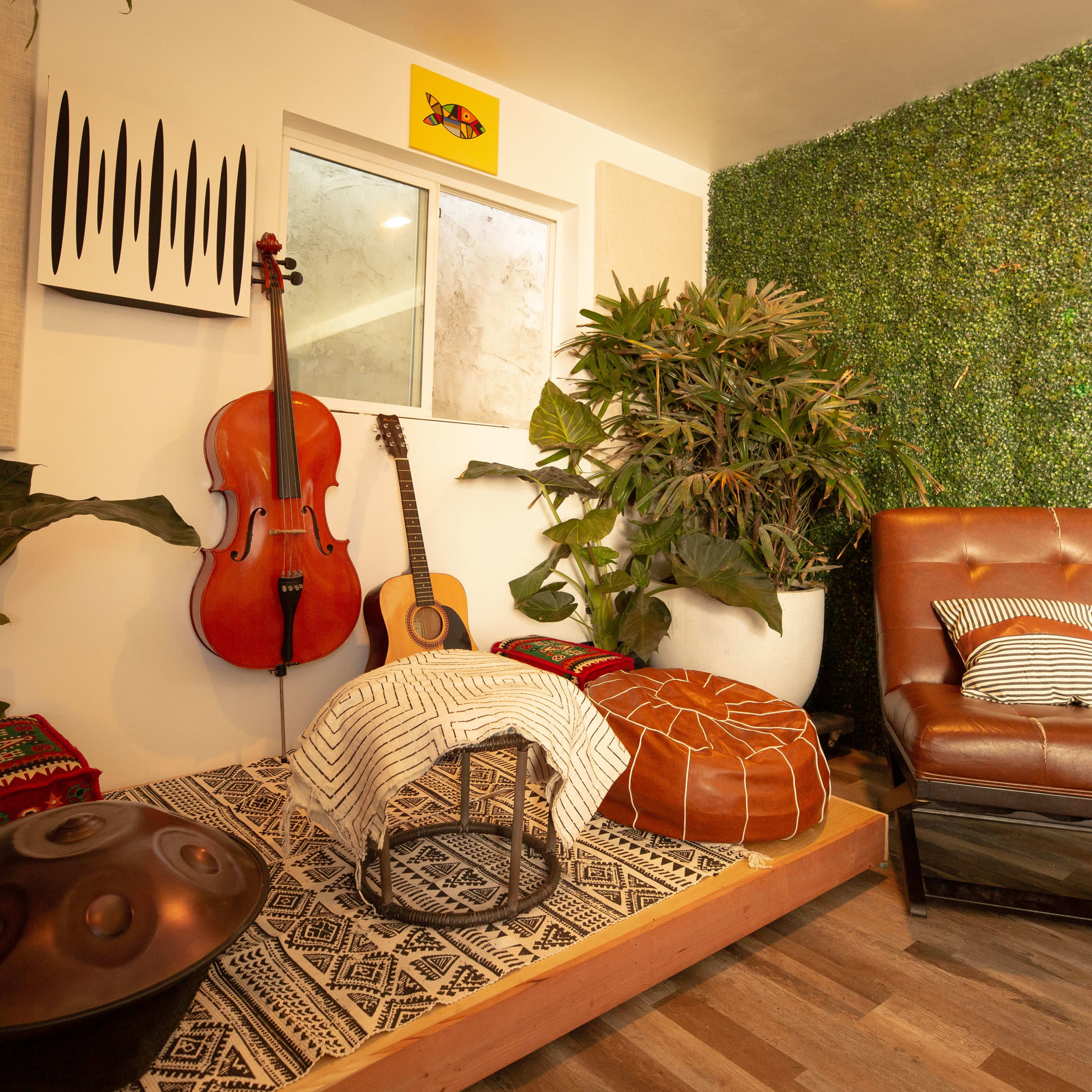 The scene features a cozy corner with a wooden platform, a brown leather chair, various musical instruments, plants, and decorative textiles against a green leafy wall.