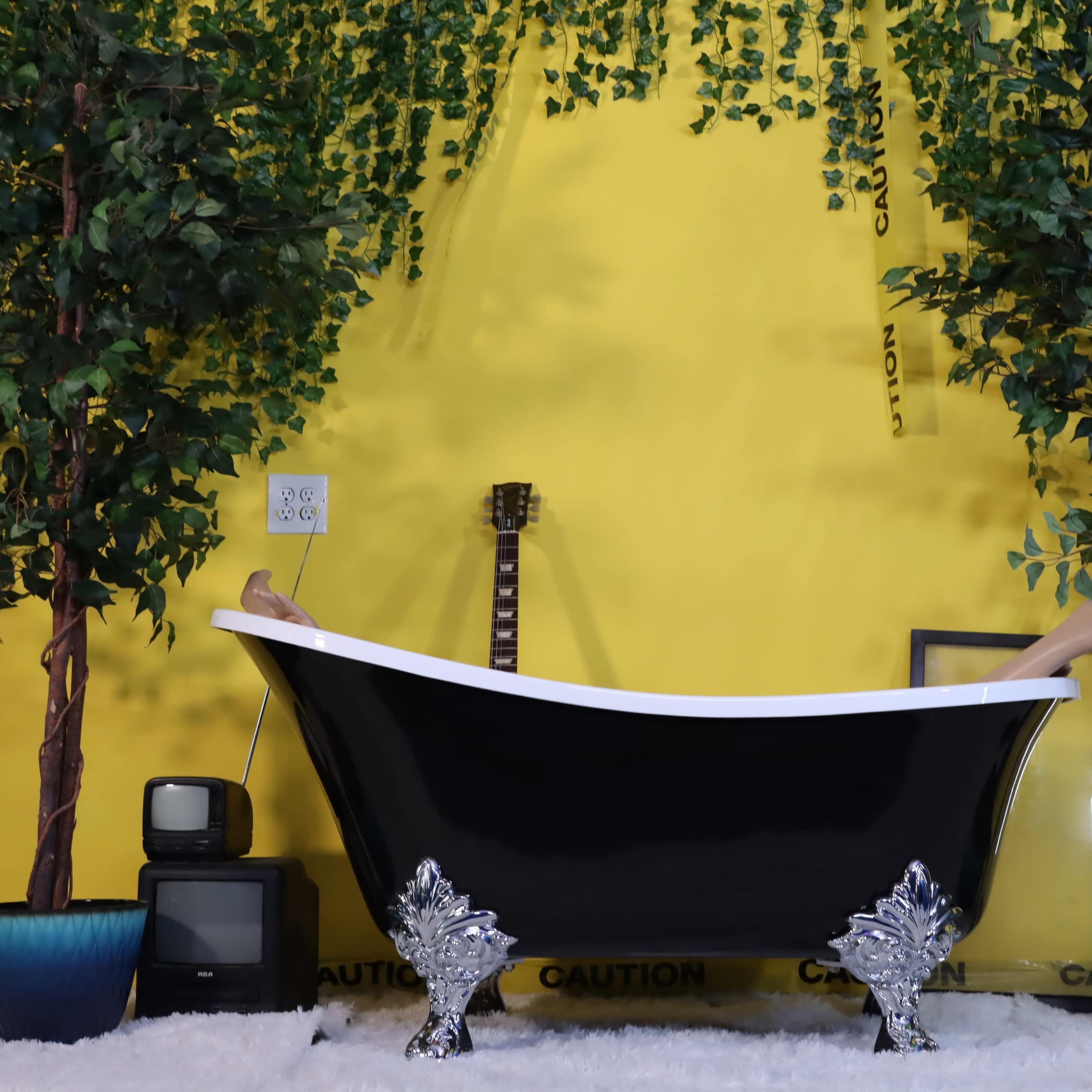 A black clawfoot bathtub is centered against a bright yellow wall, flanked by two artificial trees and a vintage television, with a guitar leaning nearby.