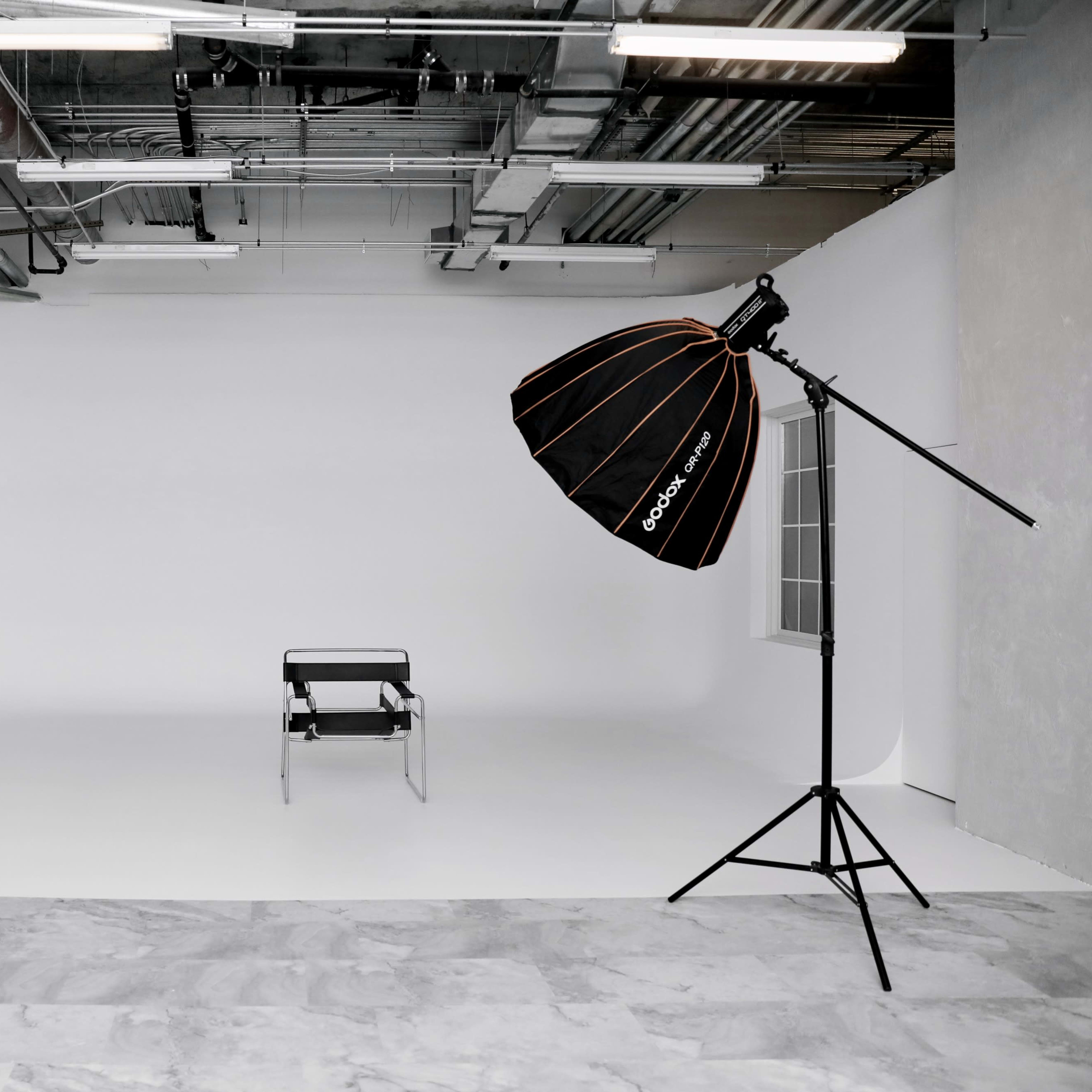 A minimalist photography studio with a single black chair and a large softbox lighting setup against a white backdrop.