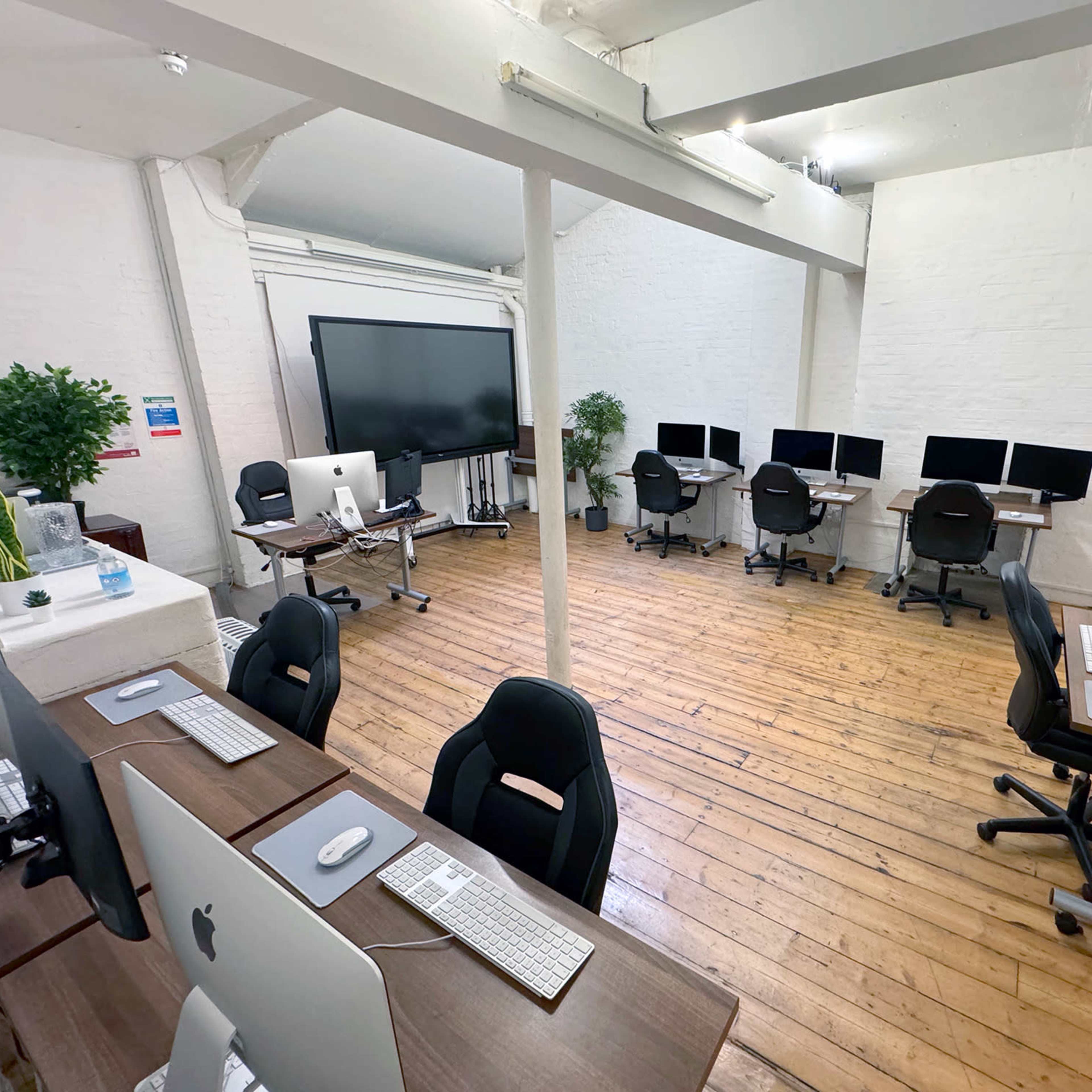 A spacious office with several desks equipped with Apple computers and ergonomic chairs, along with potted plants, arranged around a large display screen.