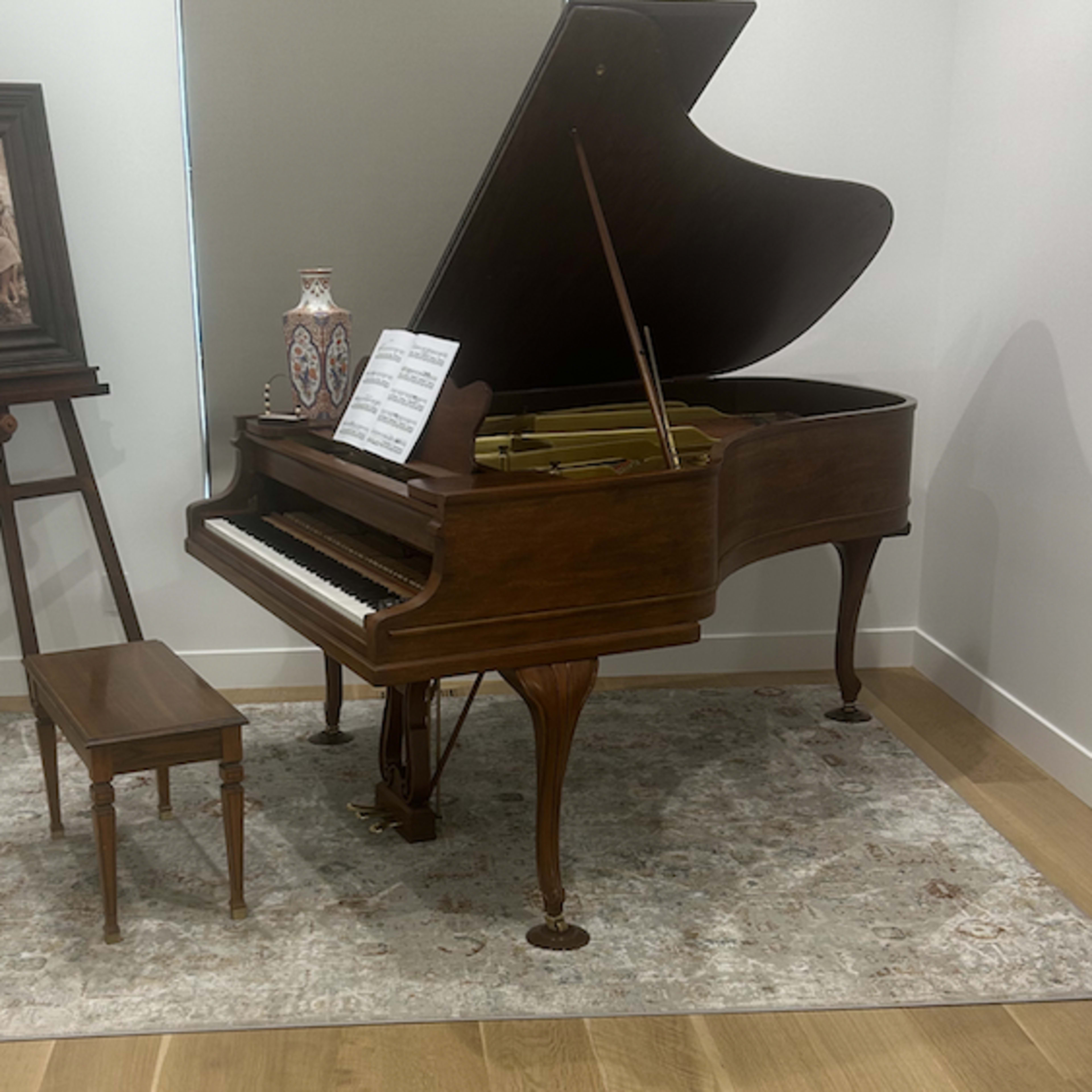 A grand piano with an open lid stands on a wooden floor near a small stool and an artwork on an easel.