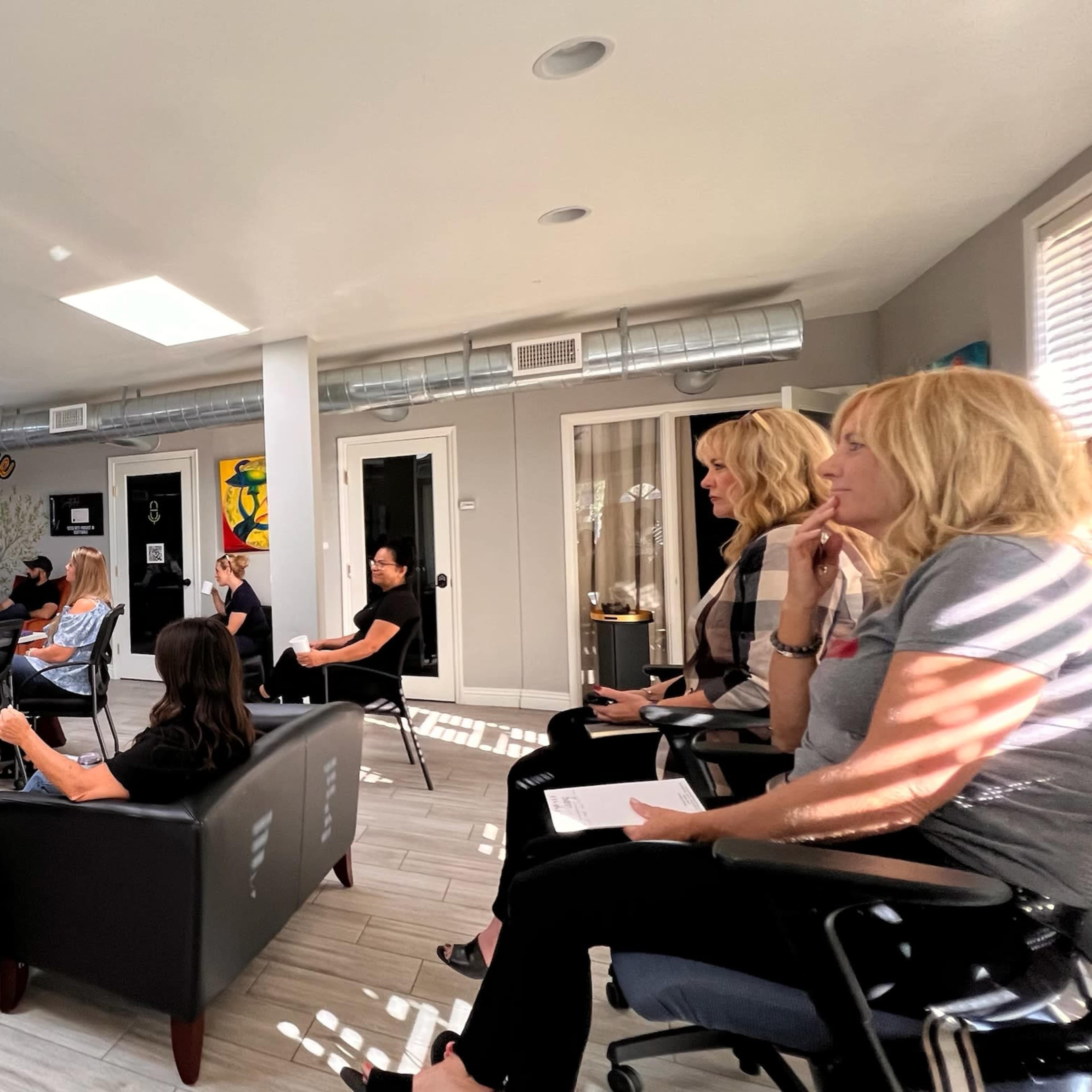 A group of adults is seated in a modern meeting space, attentively listening to a speaker presenting information on a screen.