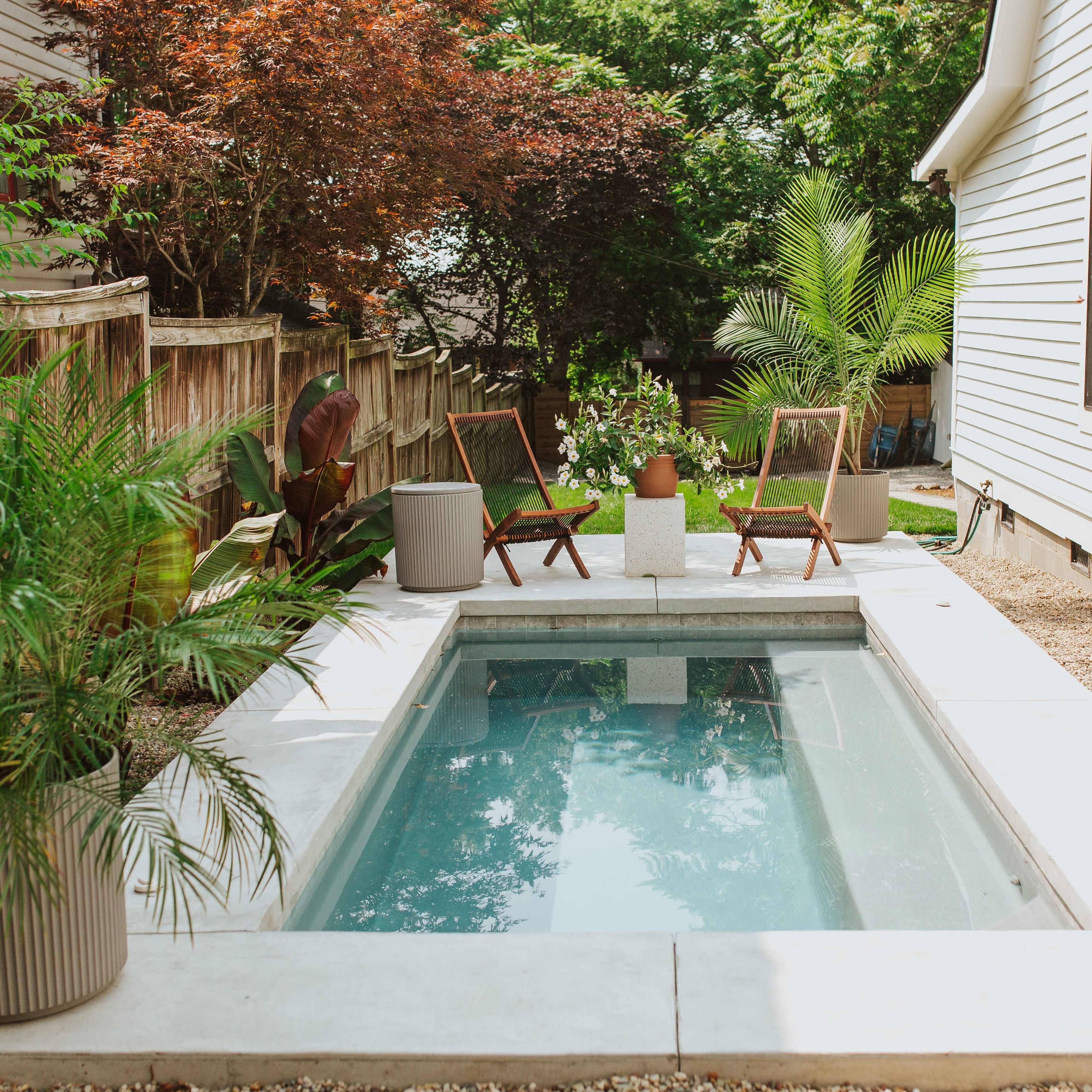 A small, modern plunge pool is surrounded by potted plants and two wooden chairs on a patio beside a house.