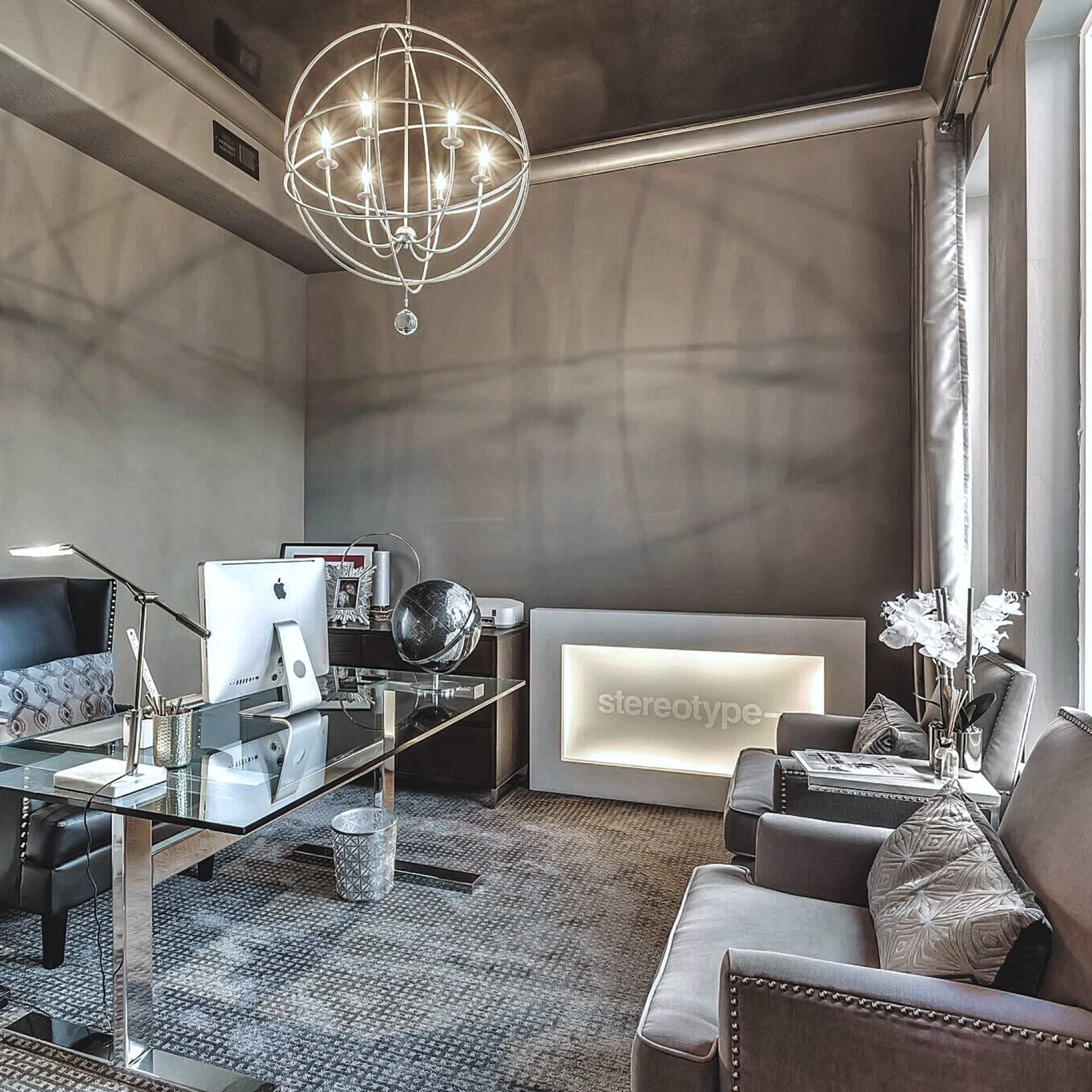 A modern office space featuring a glass desk with a computer, a black leather chair, a gray sofa, and decorative elements including a globe and a chandelier.