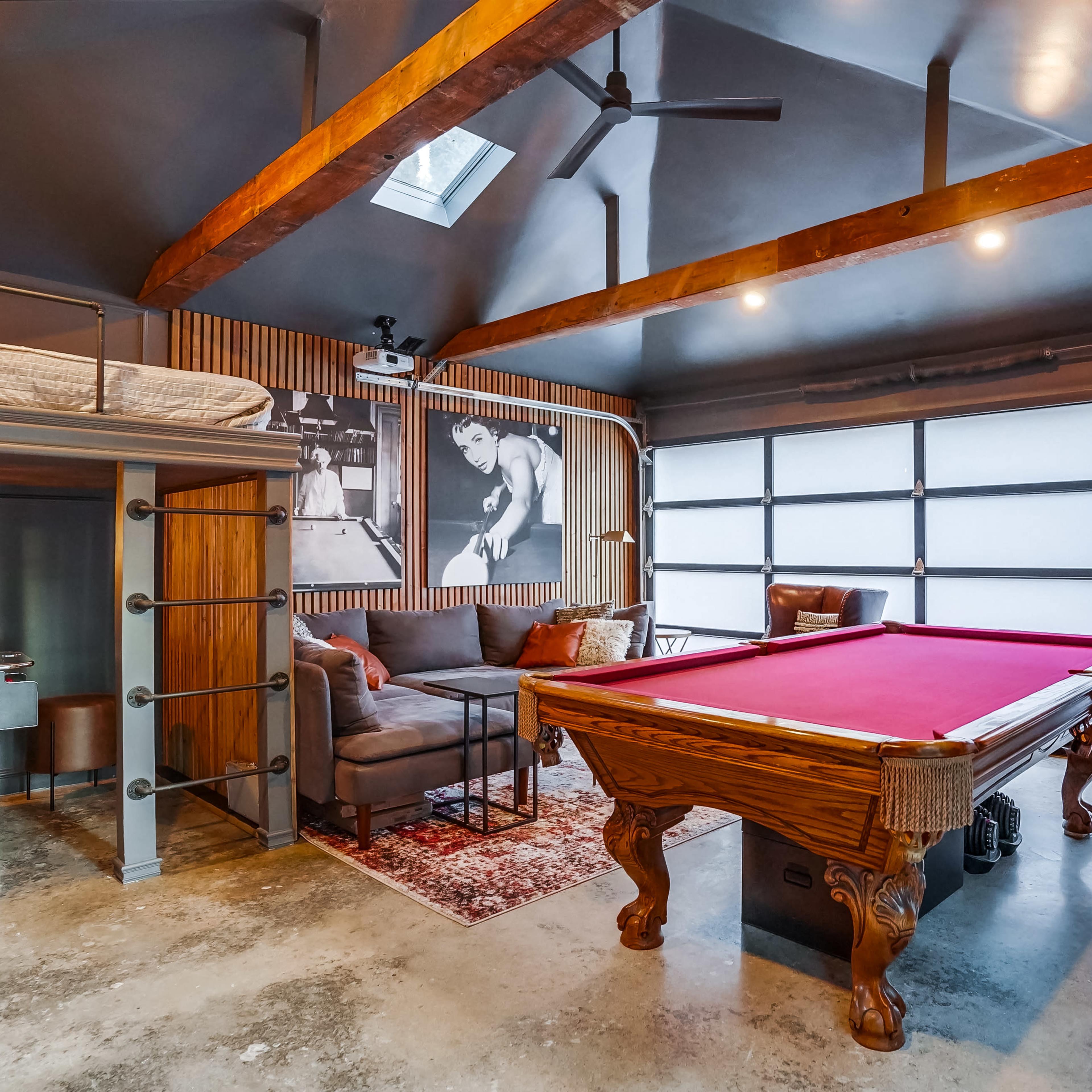 A modern game room featuring a billiards table, a seating area, and a large garage door, with wooden beams and a photo on the wall.