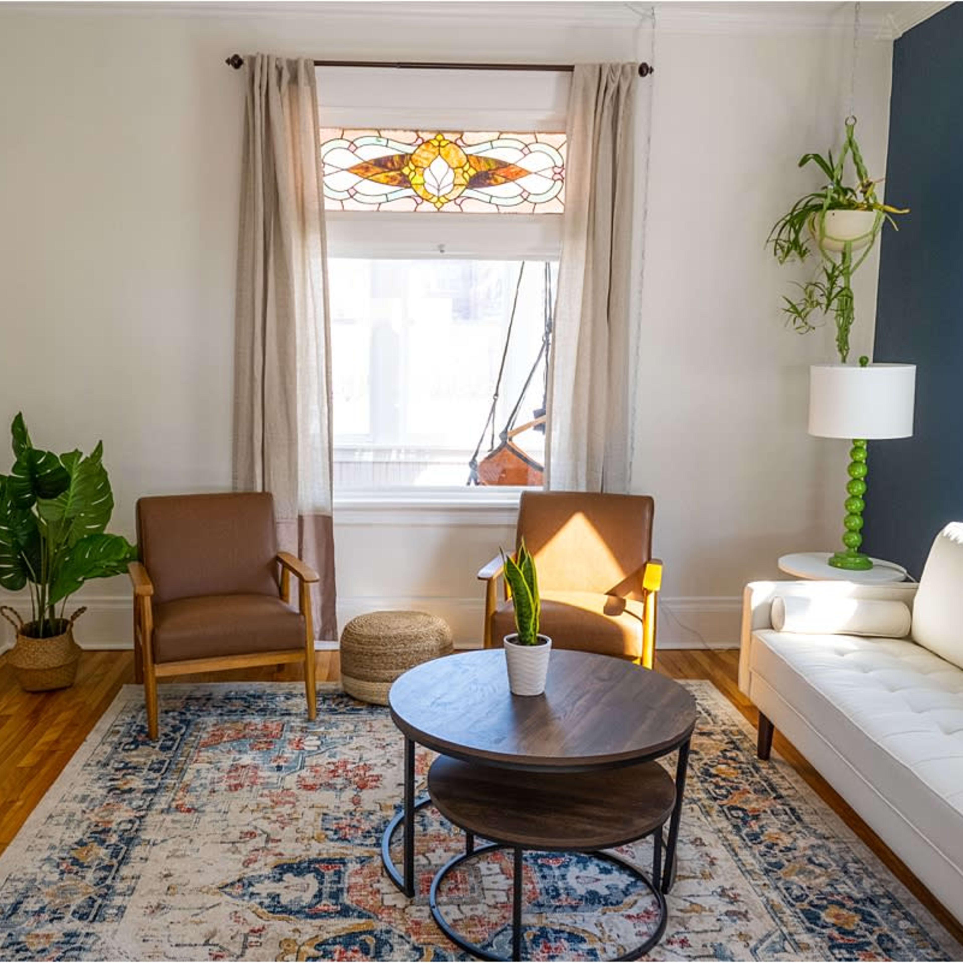 A bright living room featuring a white couch, two brown chairs, a round coffee table, a potted plant, and a lamp beside a window with a stained glass design.