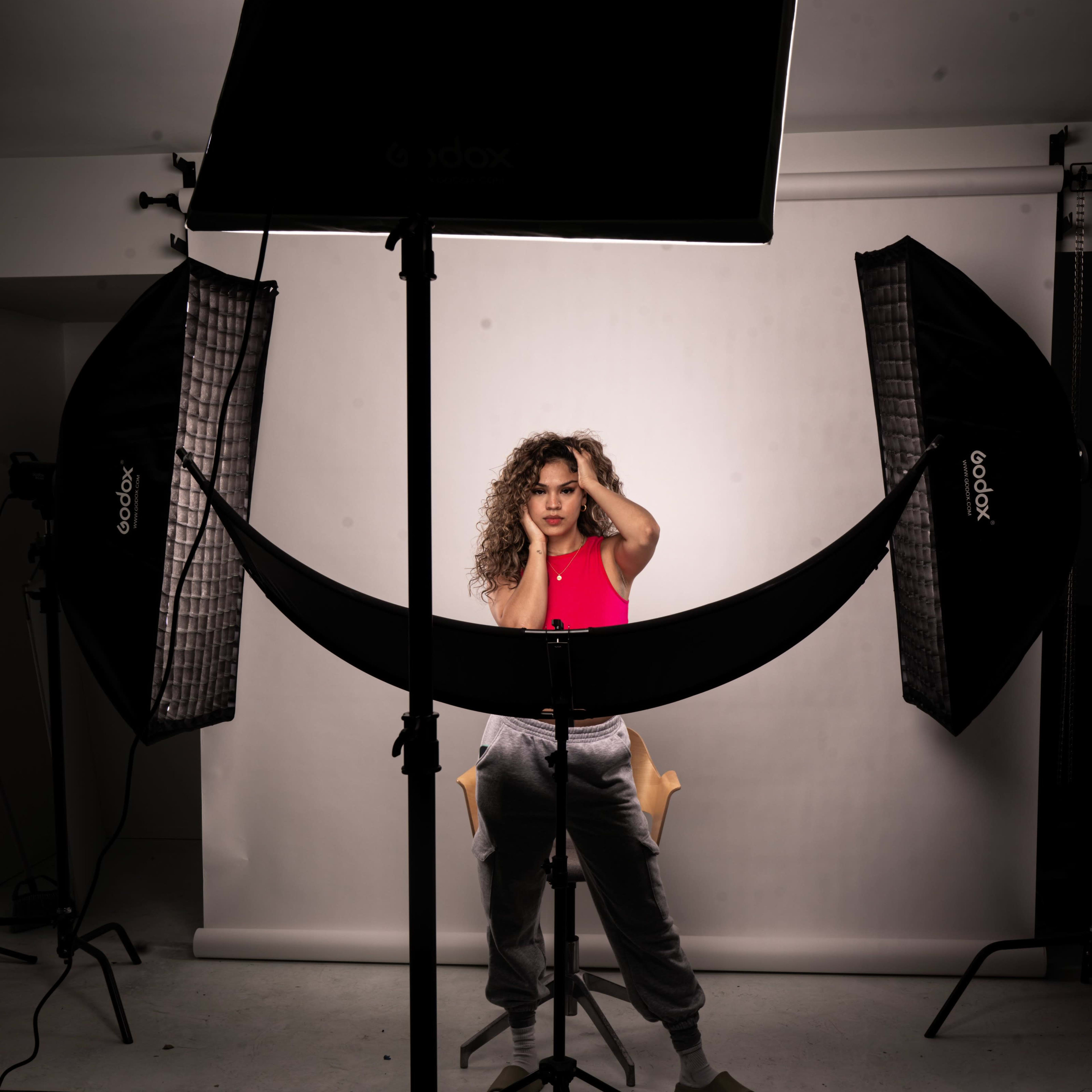 A person poses in a photography studio, surrounded by softbox lights and sitting on a stool against a white backdrop.
