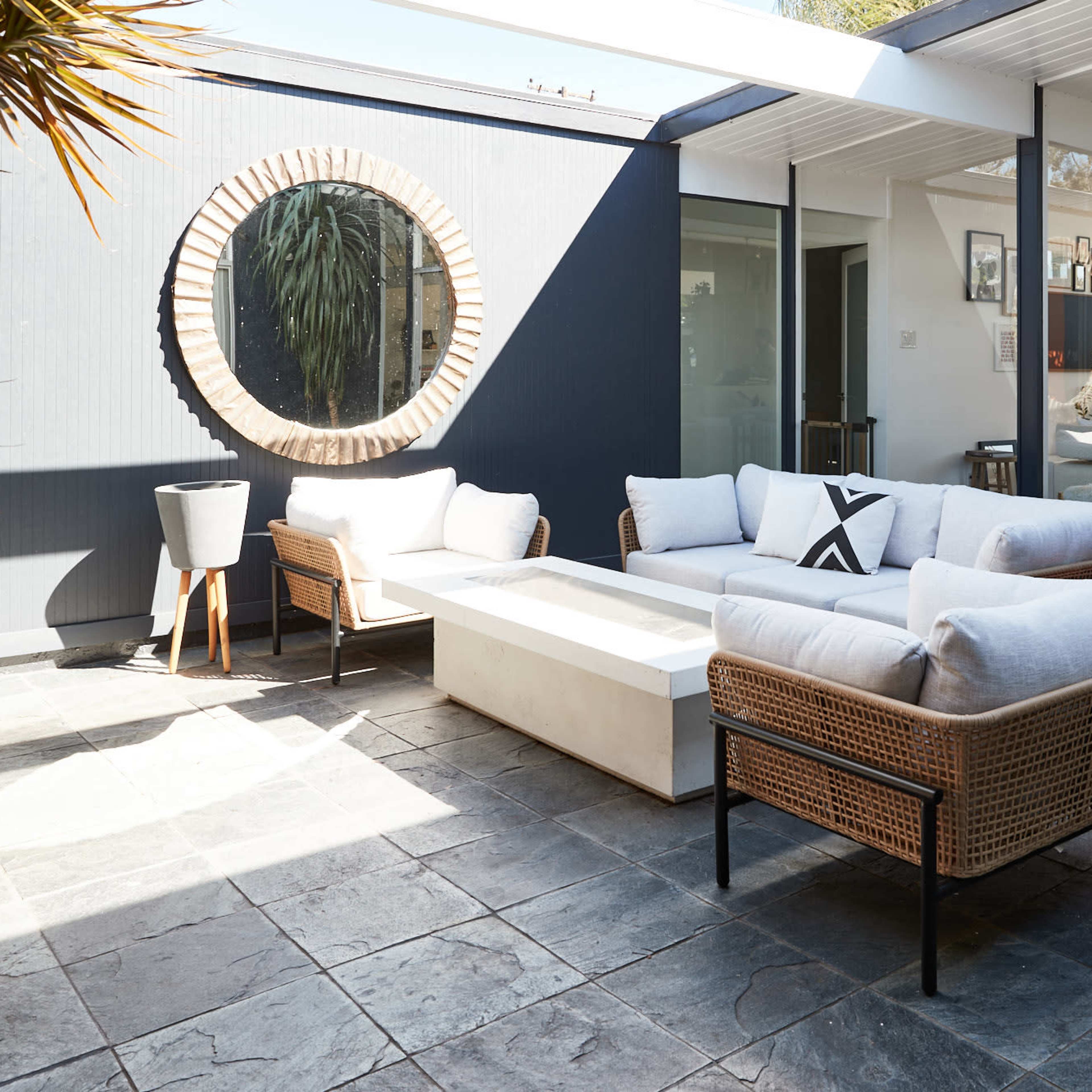 An outdoor seating area with a round mirror on a wall, featuring a white couch, wicker chairs, and a light-colored coffee table on a stone patio.