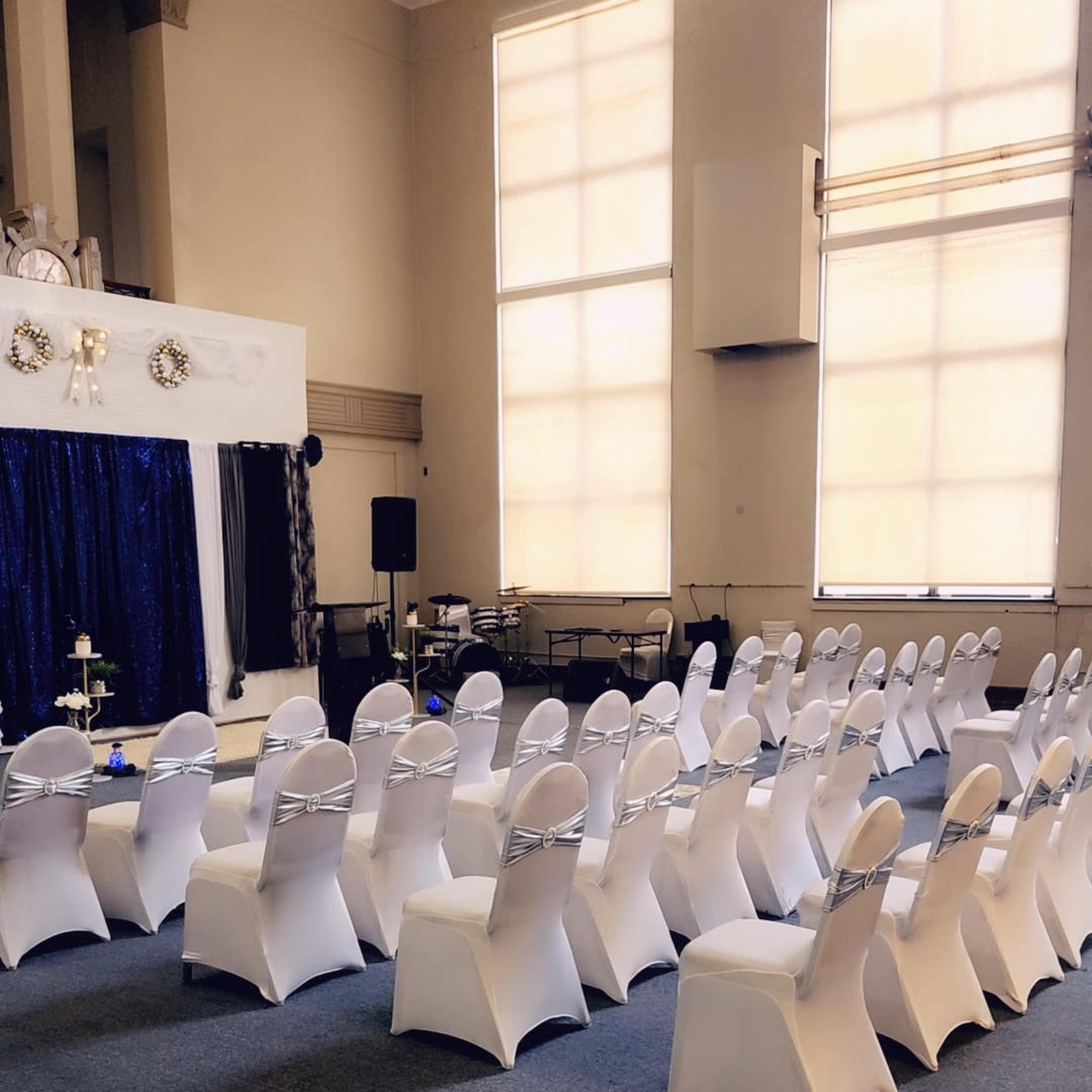 A formal event space is arranged with rows of white-covered chairs facing a decorated stage.