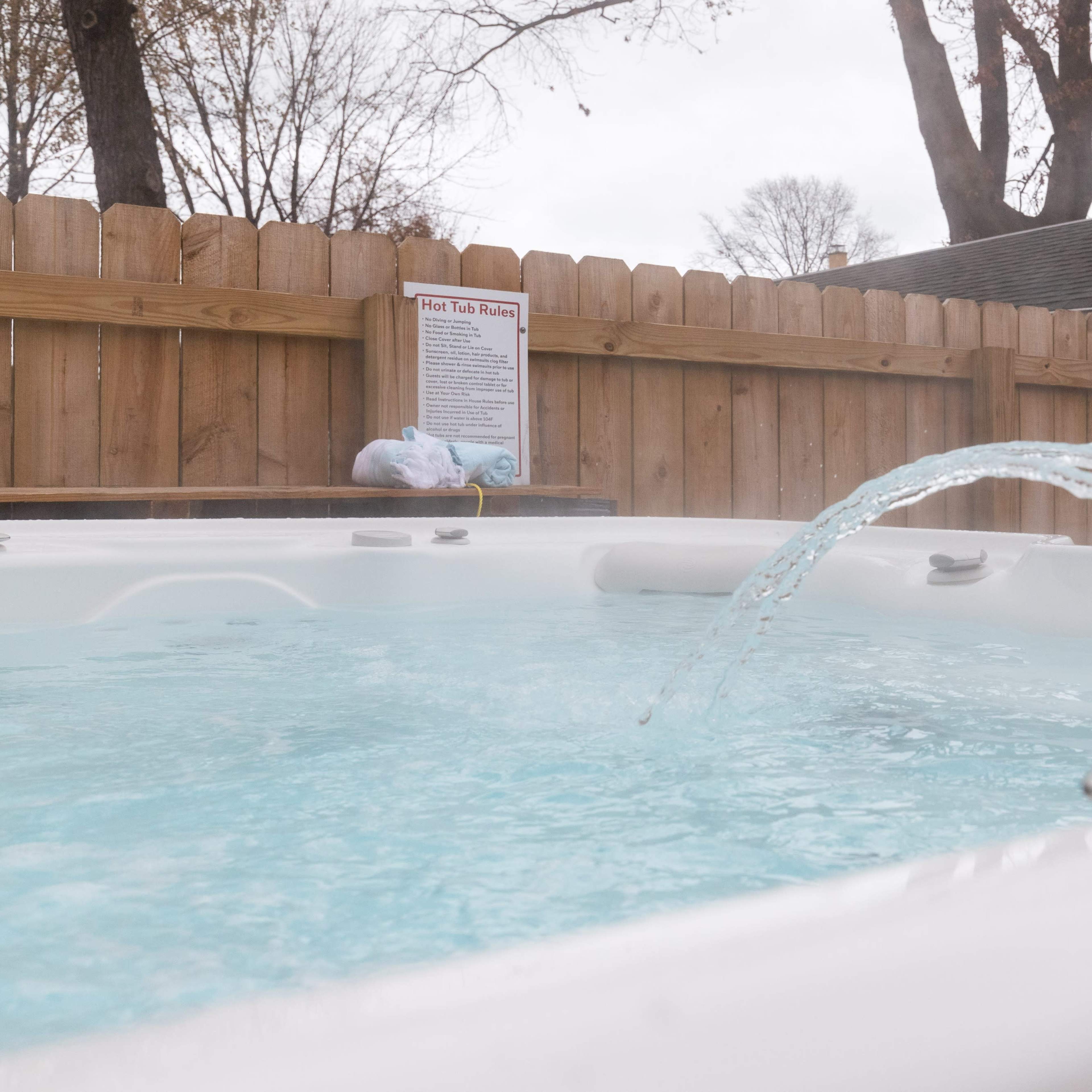 A hot tub with water jets creating arcs of water is located near a wooden fence, with a warning sign visible in the background.