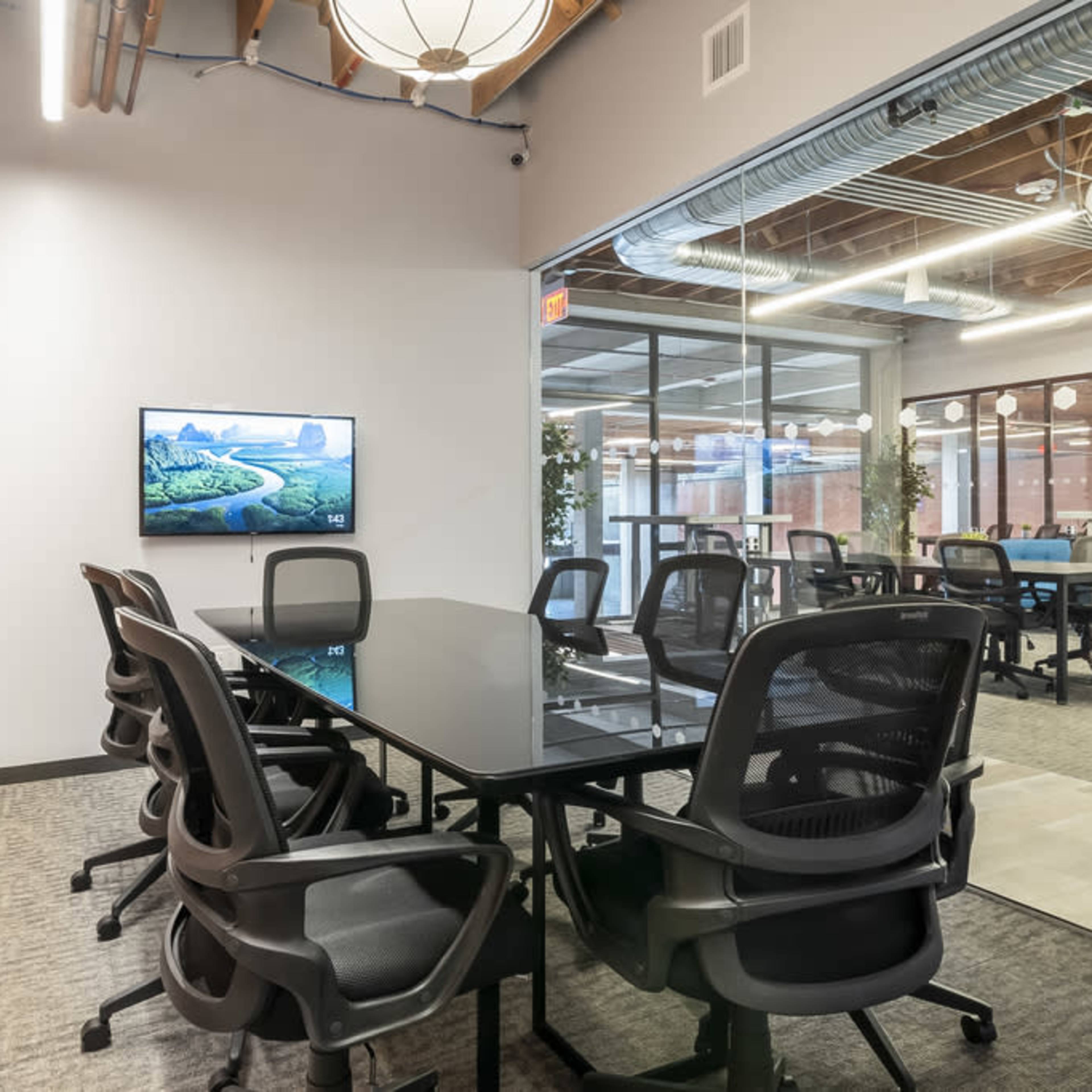 A conference room features a glass table surrounded by black chairs, with a television mounted on a brick wall.
