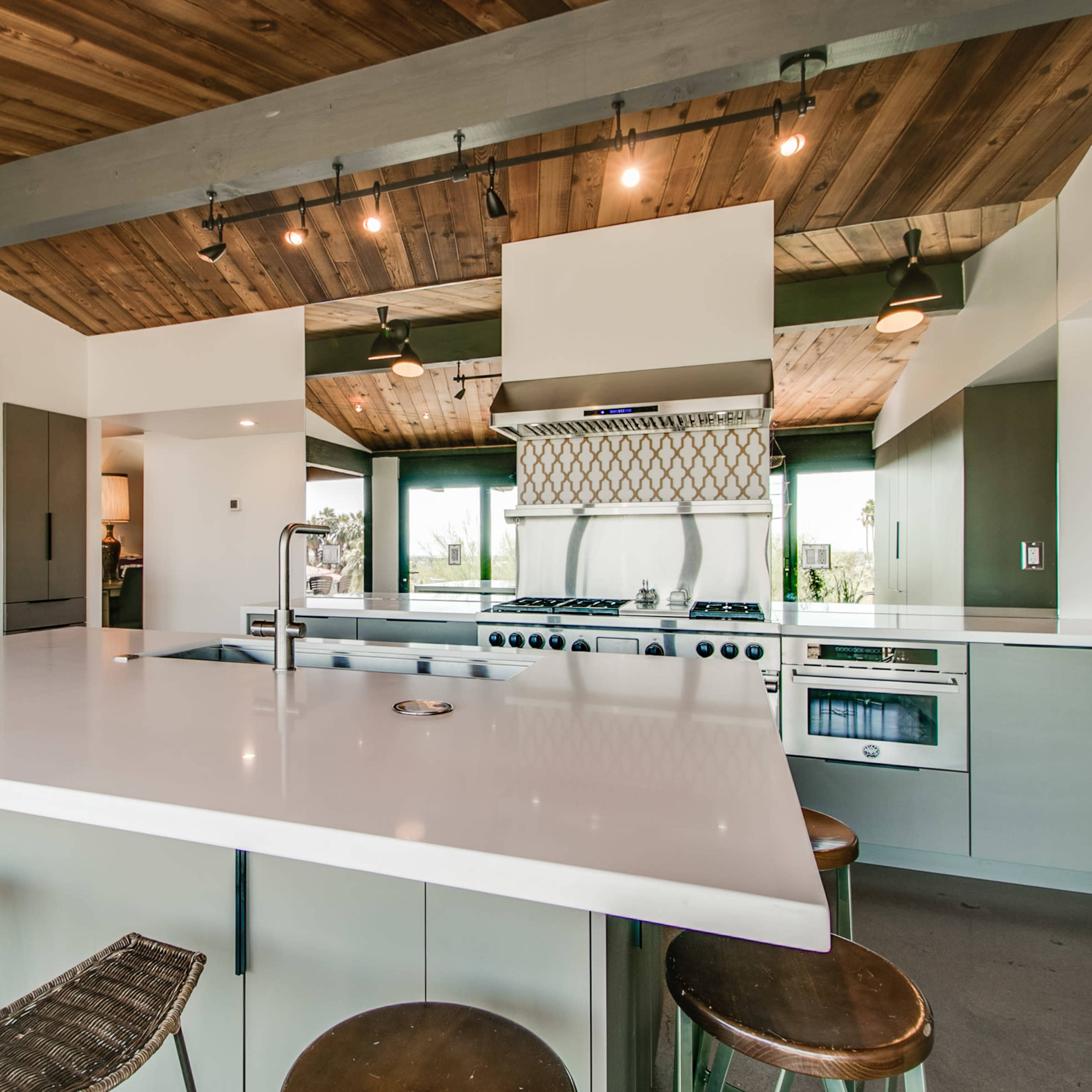 A modern kitchen features a large island with bar stools, stainless steel appliances, and wooden ceiling beams.