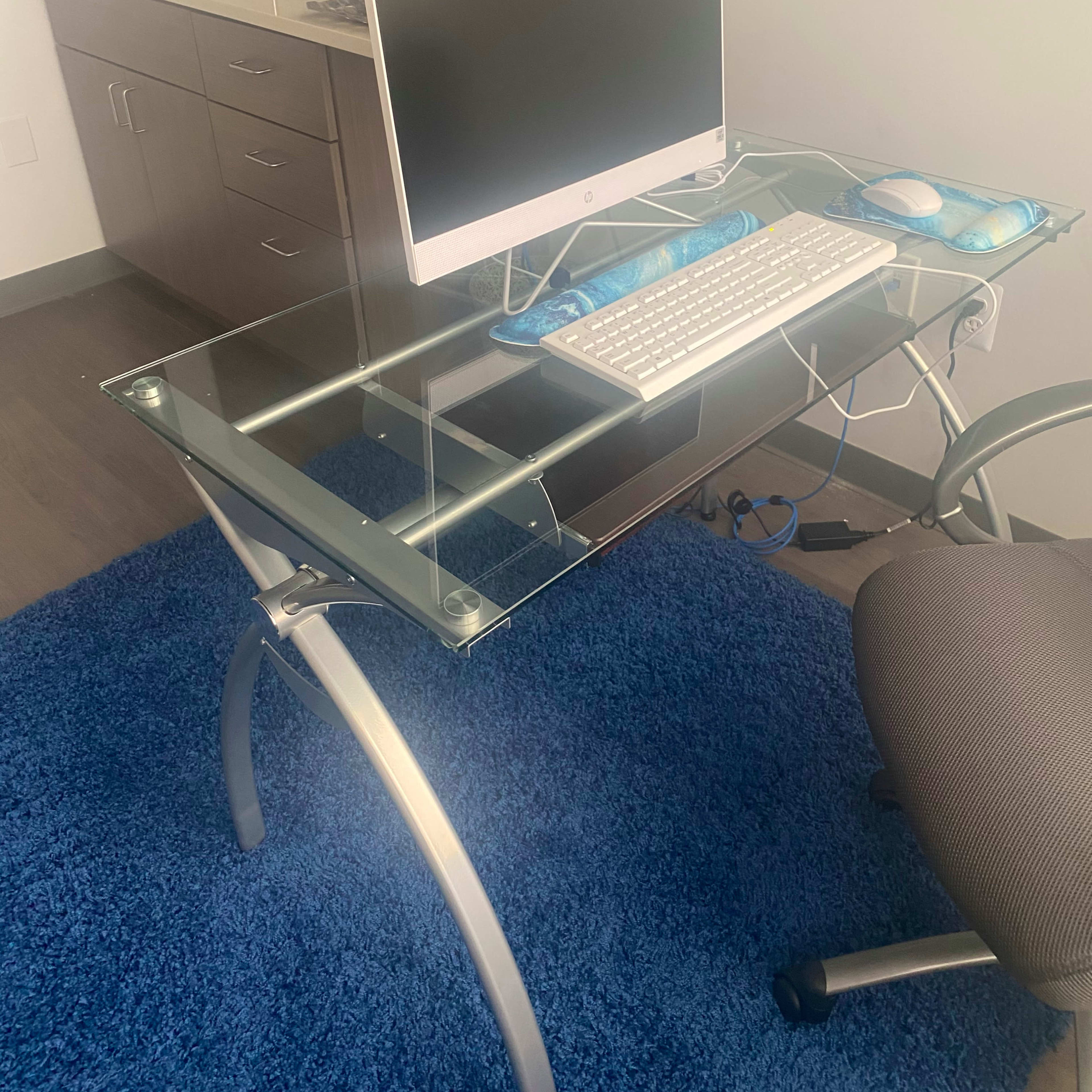 A glass computer desk with a monitor, keyboard, and mouse is positioned in front of a small kitchen area.