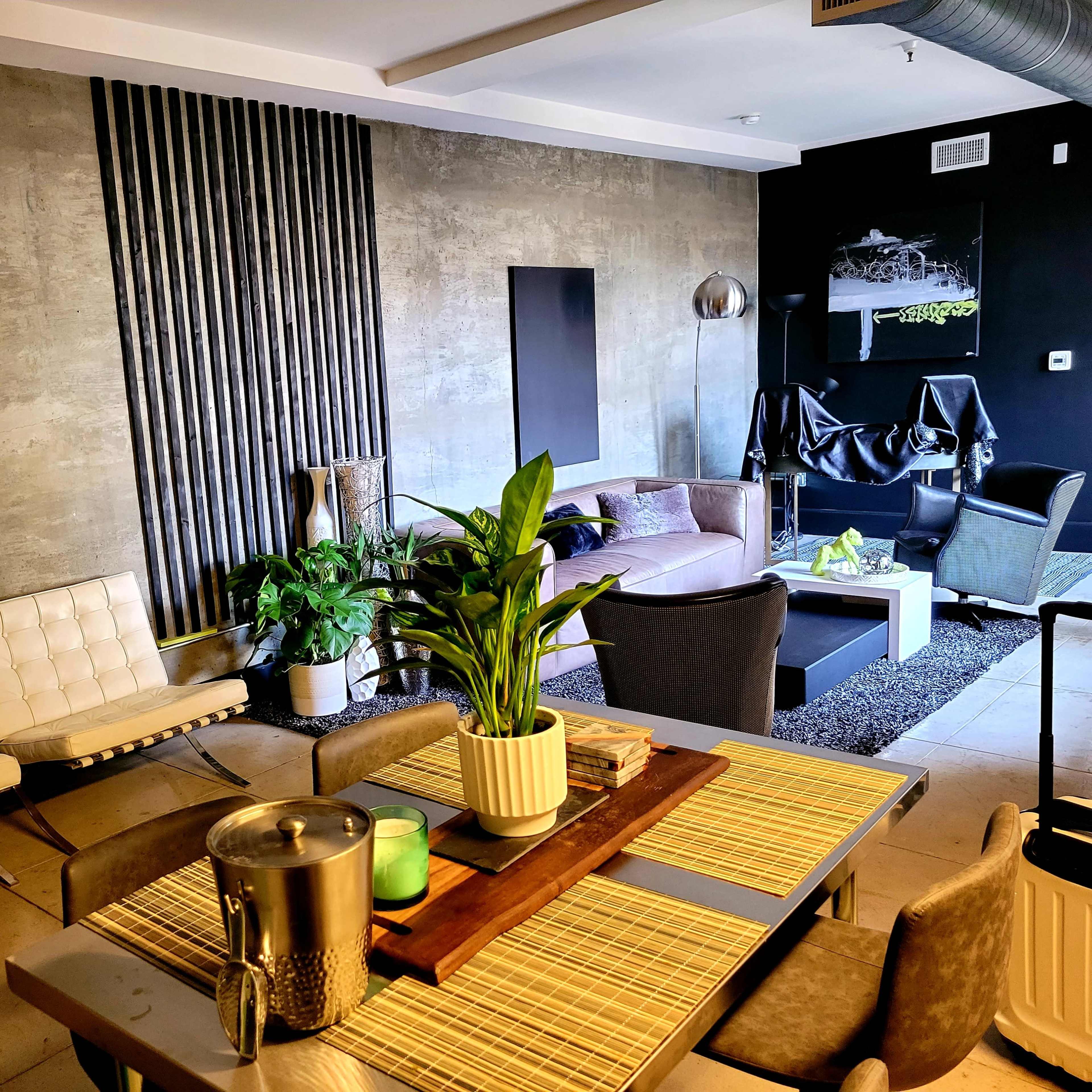 A modern living space features a dining area with a wooden table and chairs, a seating area with plush couches, and decorative plants against a concrete wall.