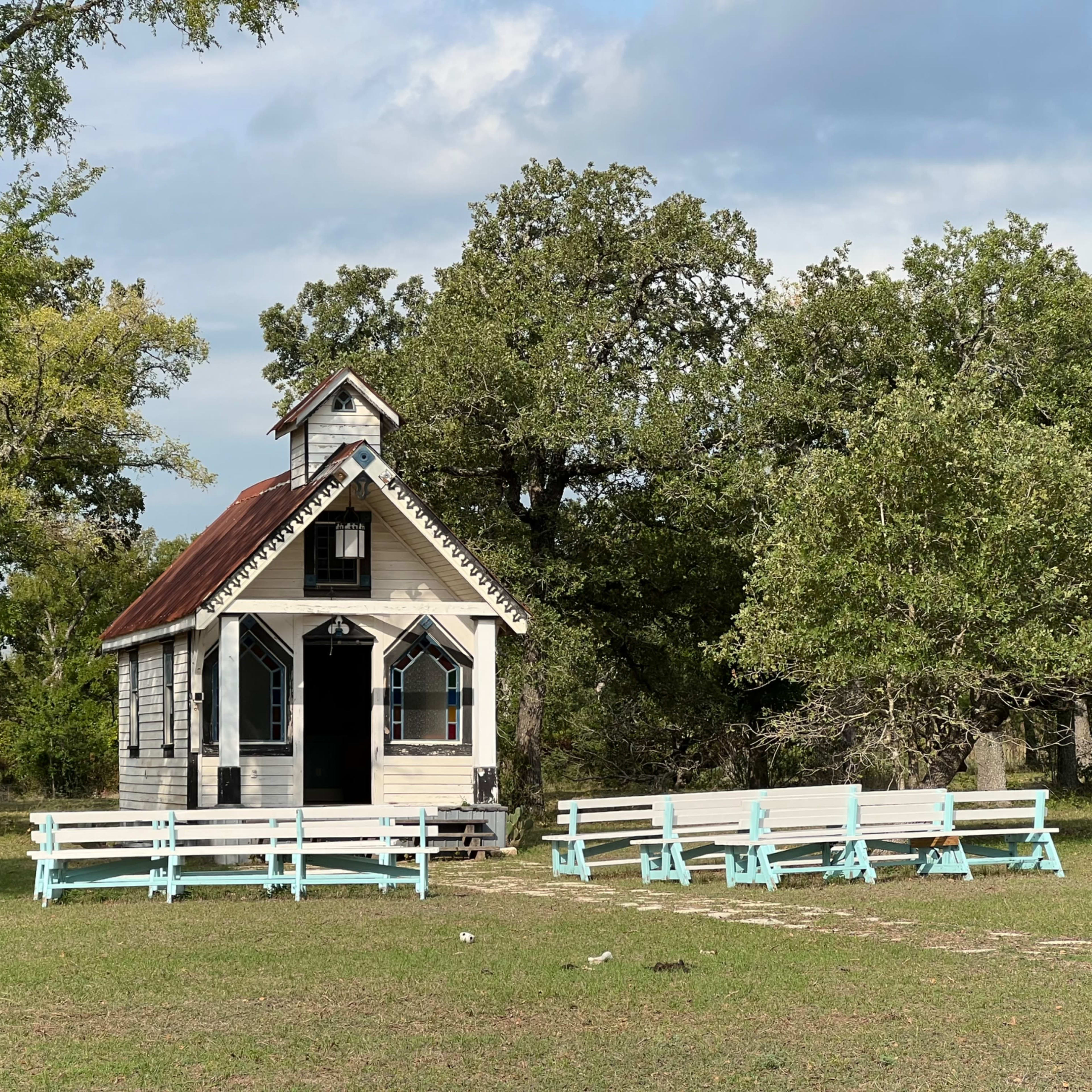 A small, white wooden chapel with a red roof sits among green trees, surrounded by several light blue benches arranged in front of it.