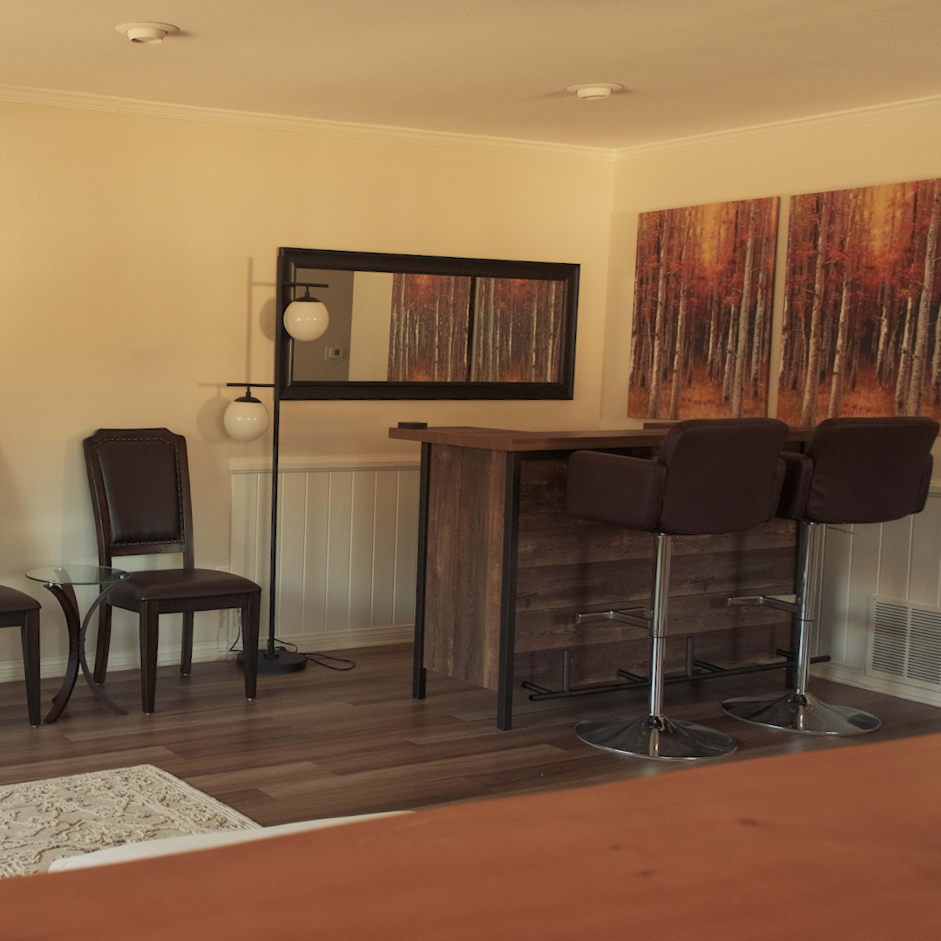 A living room features a large television, two chairs, a bar with stools, and a decorative painting of trees on the wall.