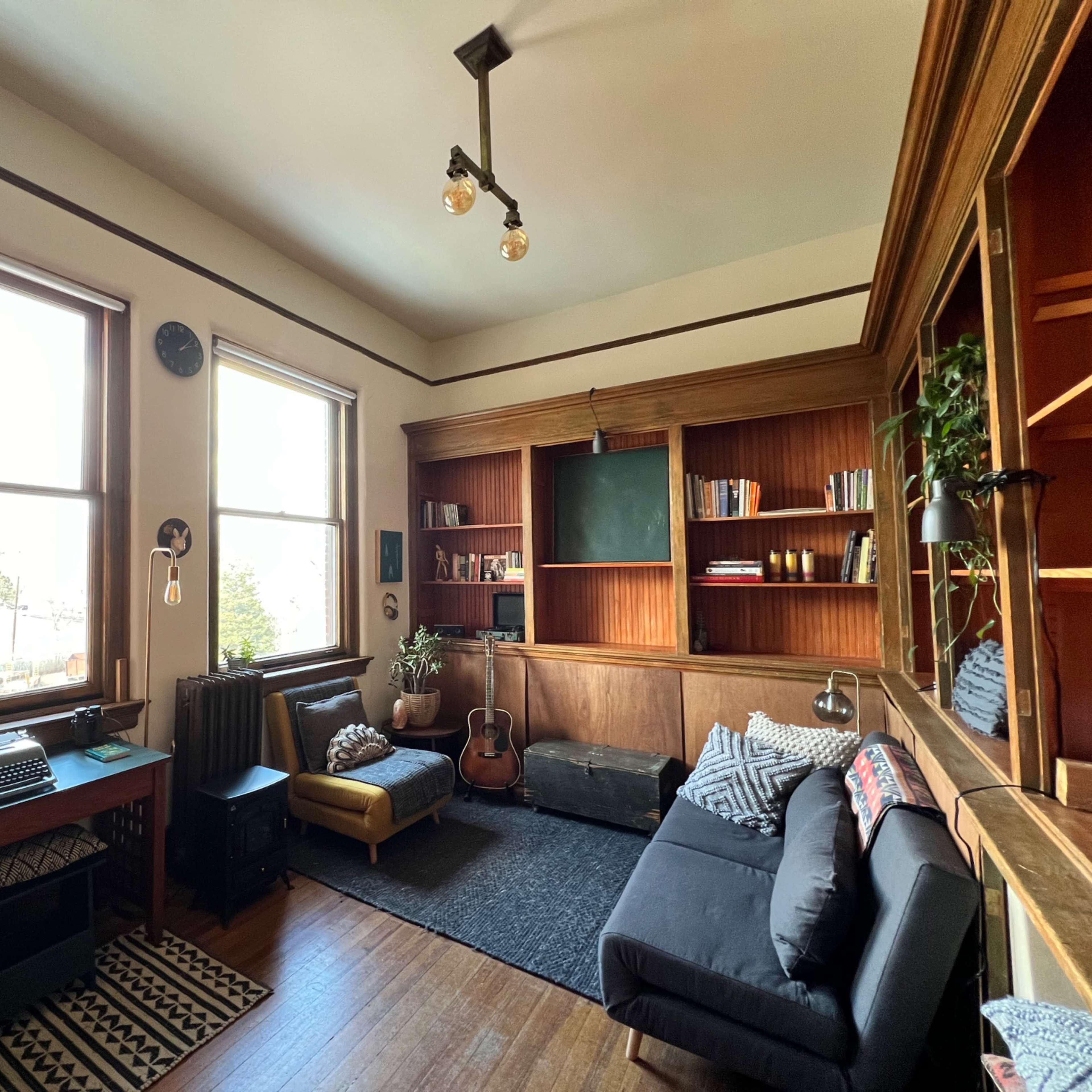 A cozy room featuring large windows, a wooden bookshelf, a black sofa, a vintage typewriter on a desk, and a guitar leaning against the wall.