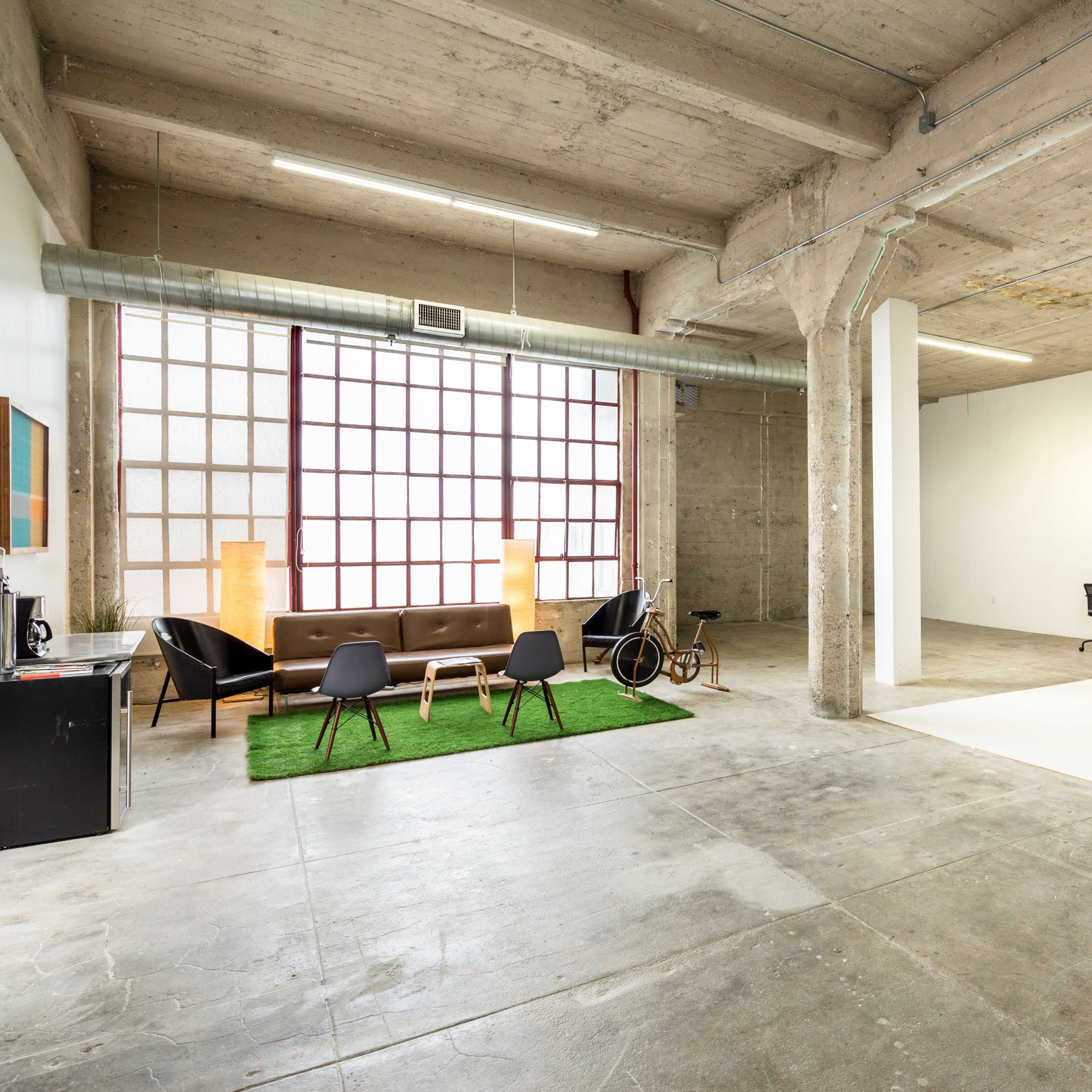 An industrial-style interior space with a seating area featuring a coffee table and green carpet, alongside a minimalistic workspace and a large window.