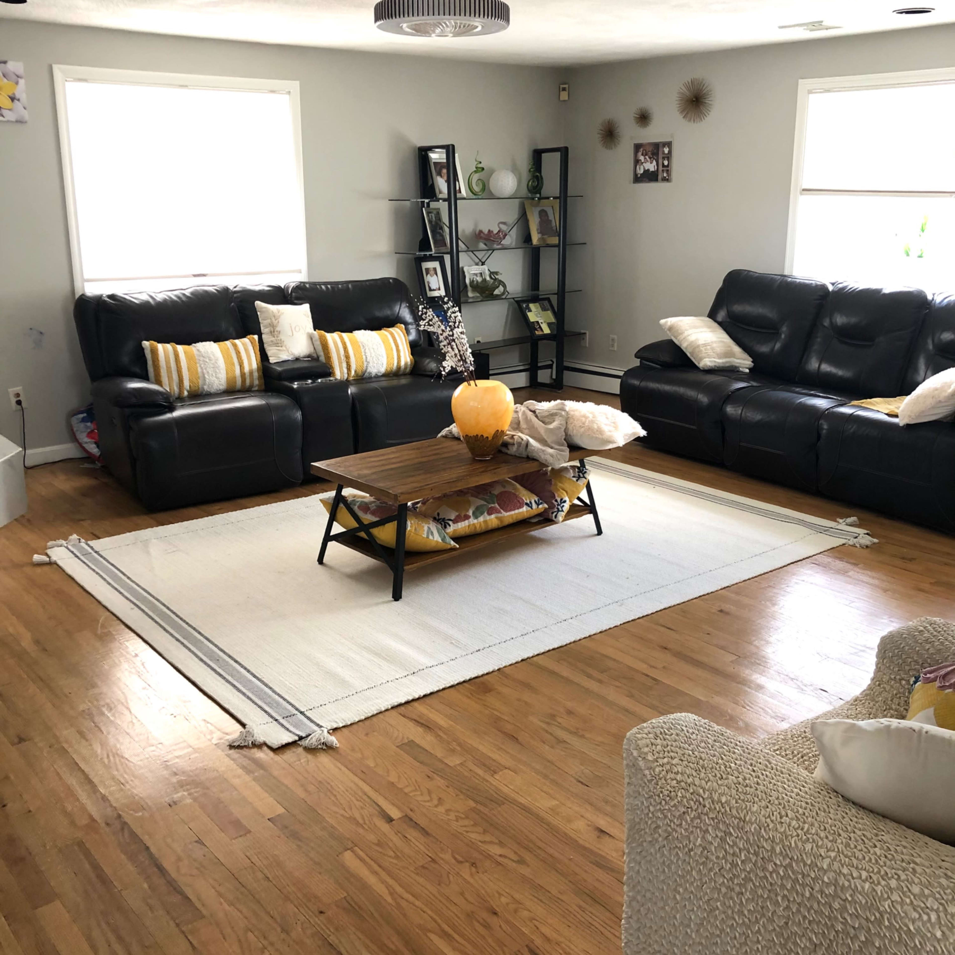 A living room with dark leather sofas, a coffee table on a rug, and a shelf displaying decorative items against light gray walls.