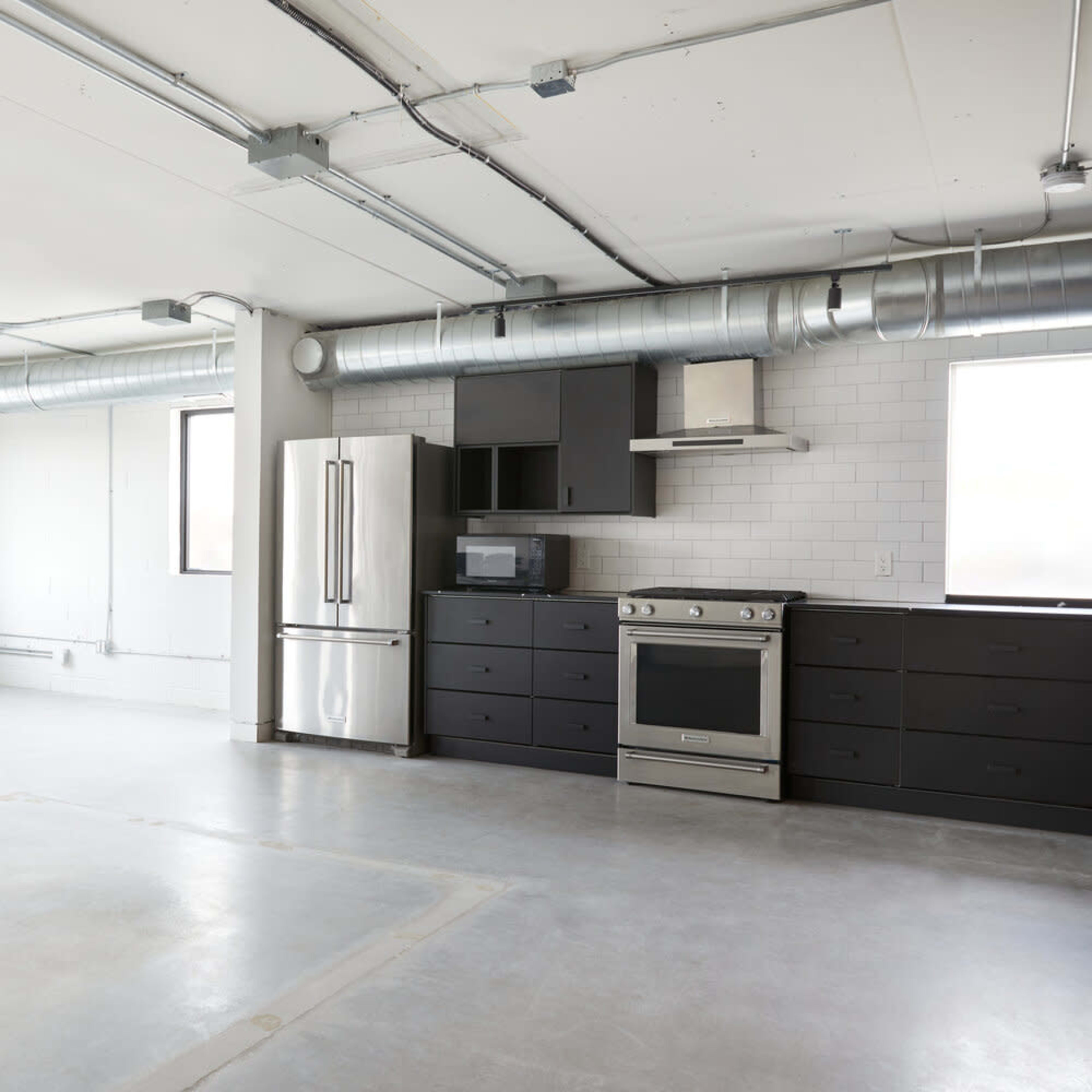A spacious, modern kitchen area with stainless steel appliances and concrete flooring, featuring large windows on two walls.