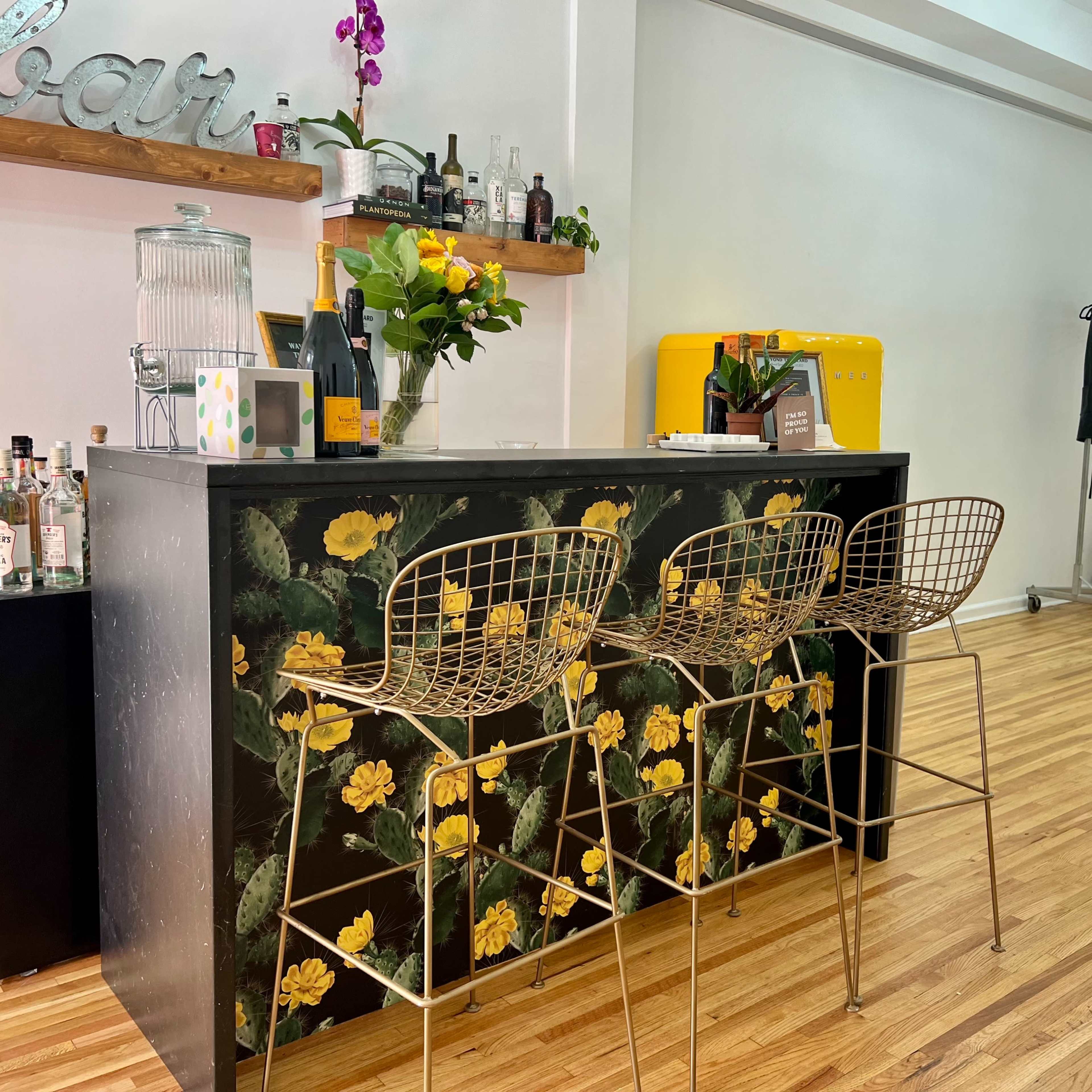 A bar with a floral design is positioned against a wall, featuring three gold wire bar stools and a yellow refrigerator in the background.