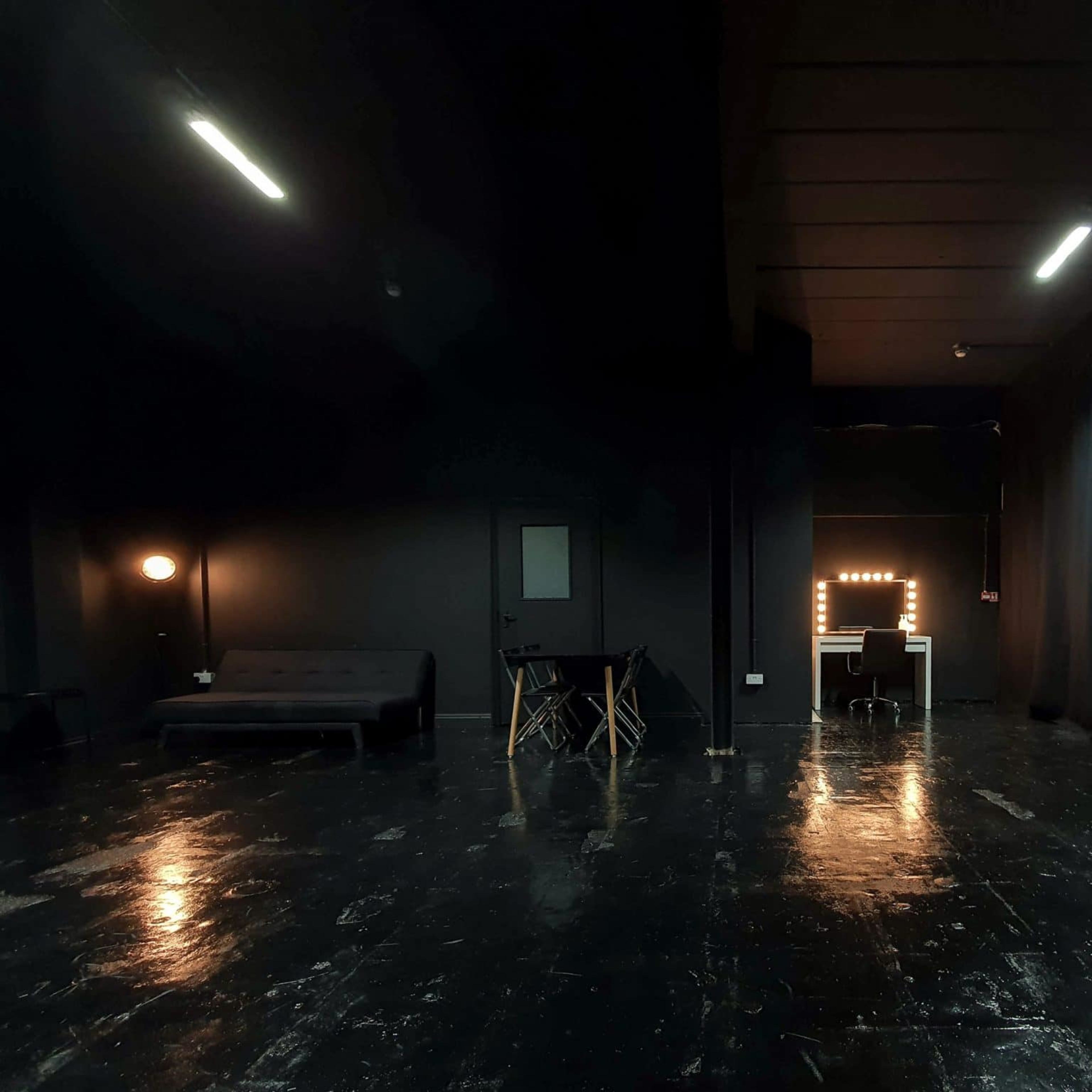 A dimly lit room with dark walls, a sofa on the left, a table and chairs in the center, and a brightly lit vanity mirror against the back wall.