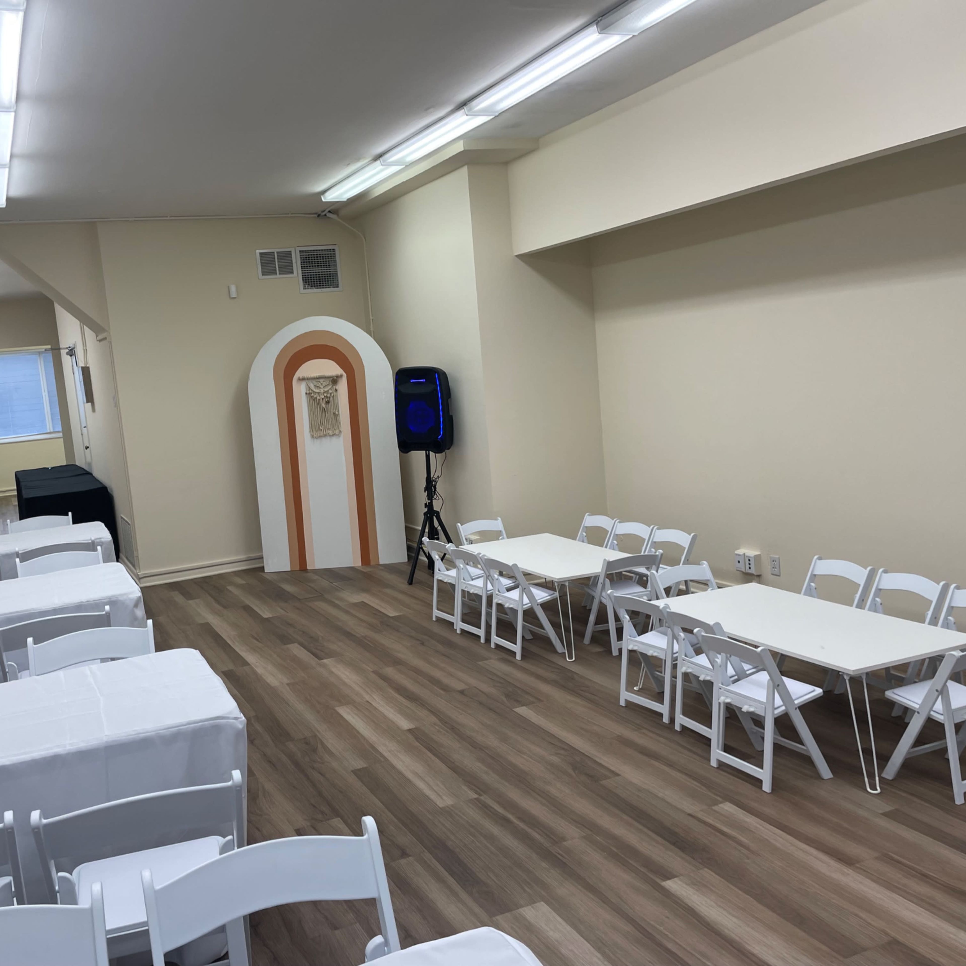 A spacious room with white folding chairs and tables arranged on a wooden floor, illuminated by fluorescent lights.