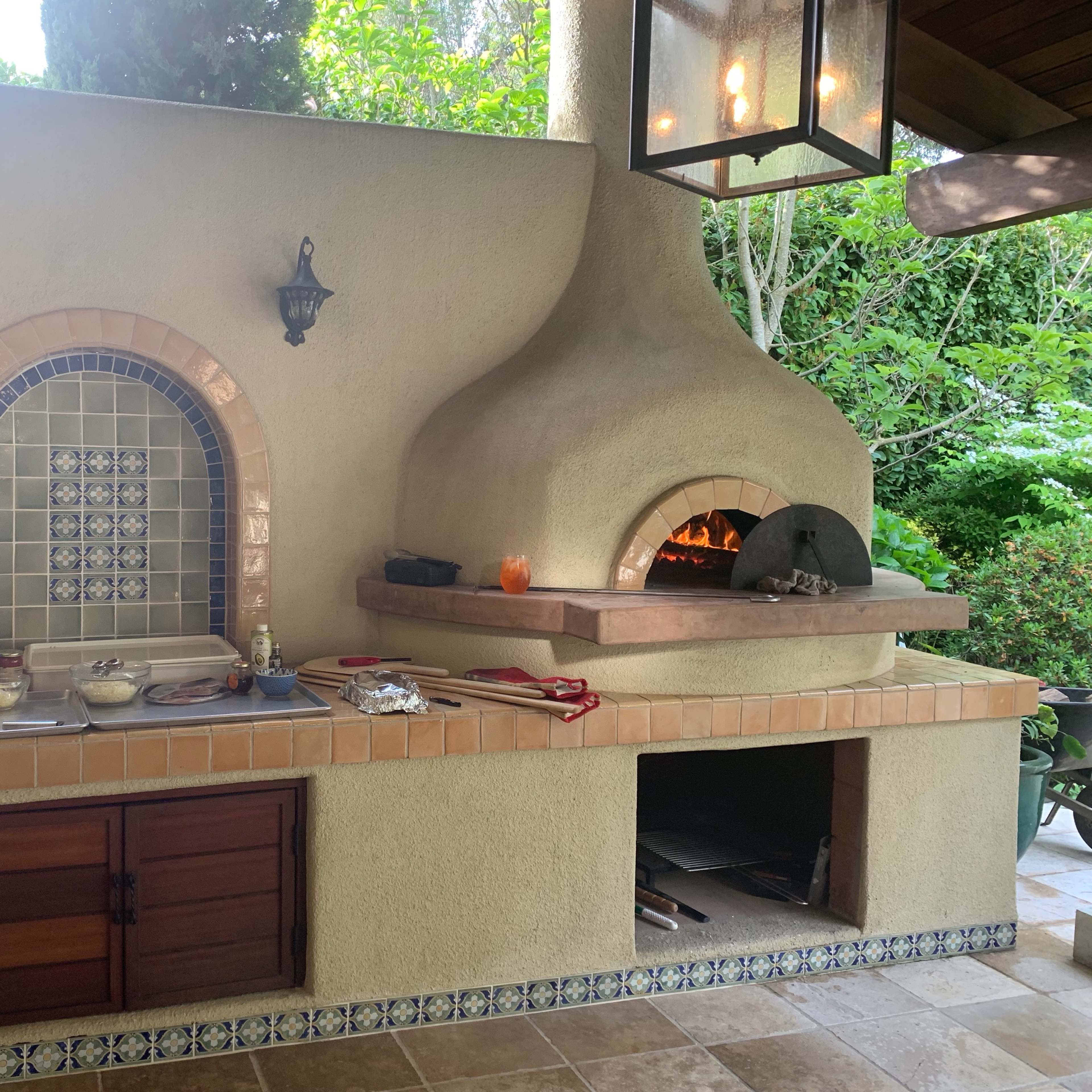 An outdoor kitchen with a stone pizza oven, a countertop for food preparation, and various cooking utensils arranged nearby.