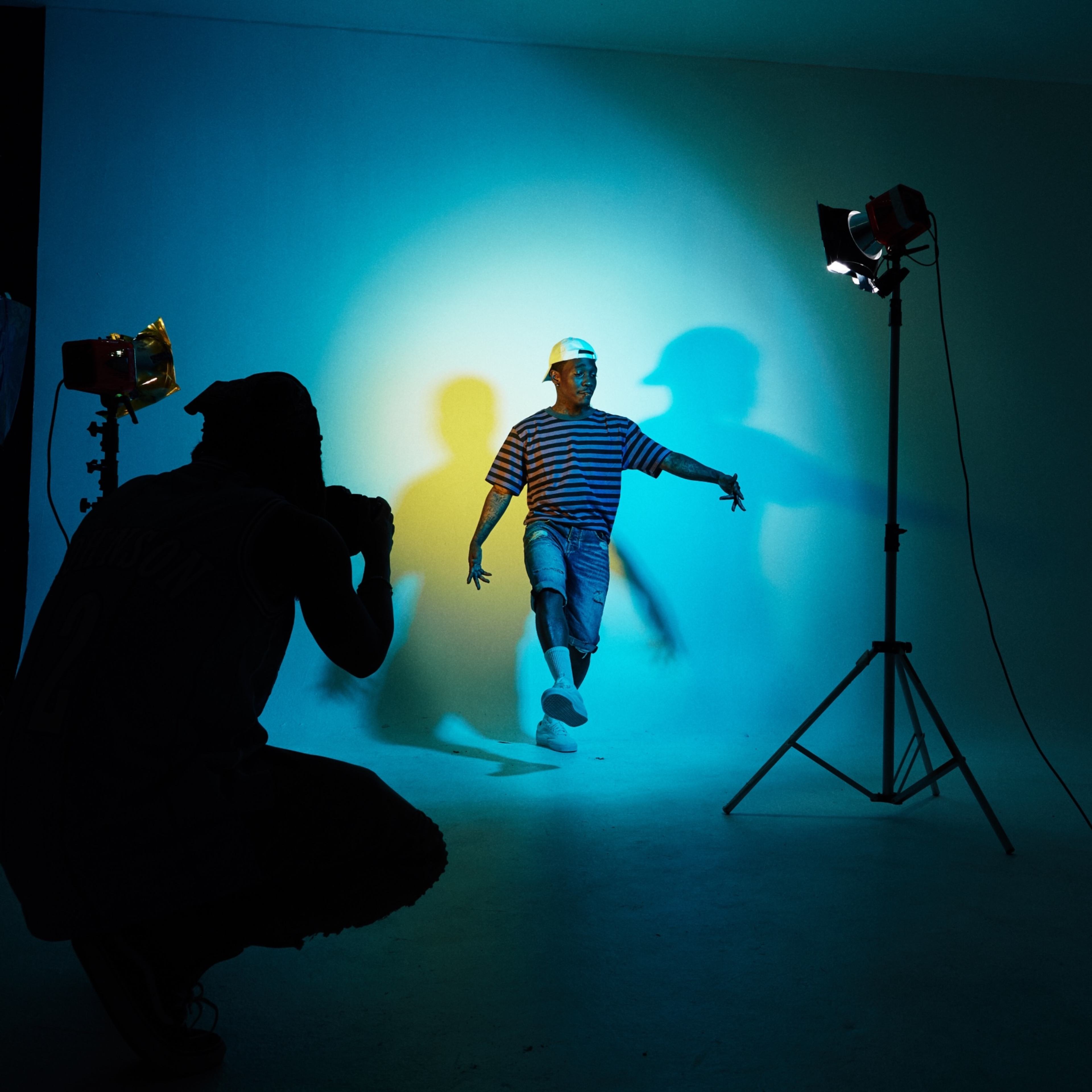 A person poses in front of a backdrop while another individual photographs them using studio lighting.