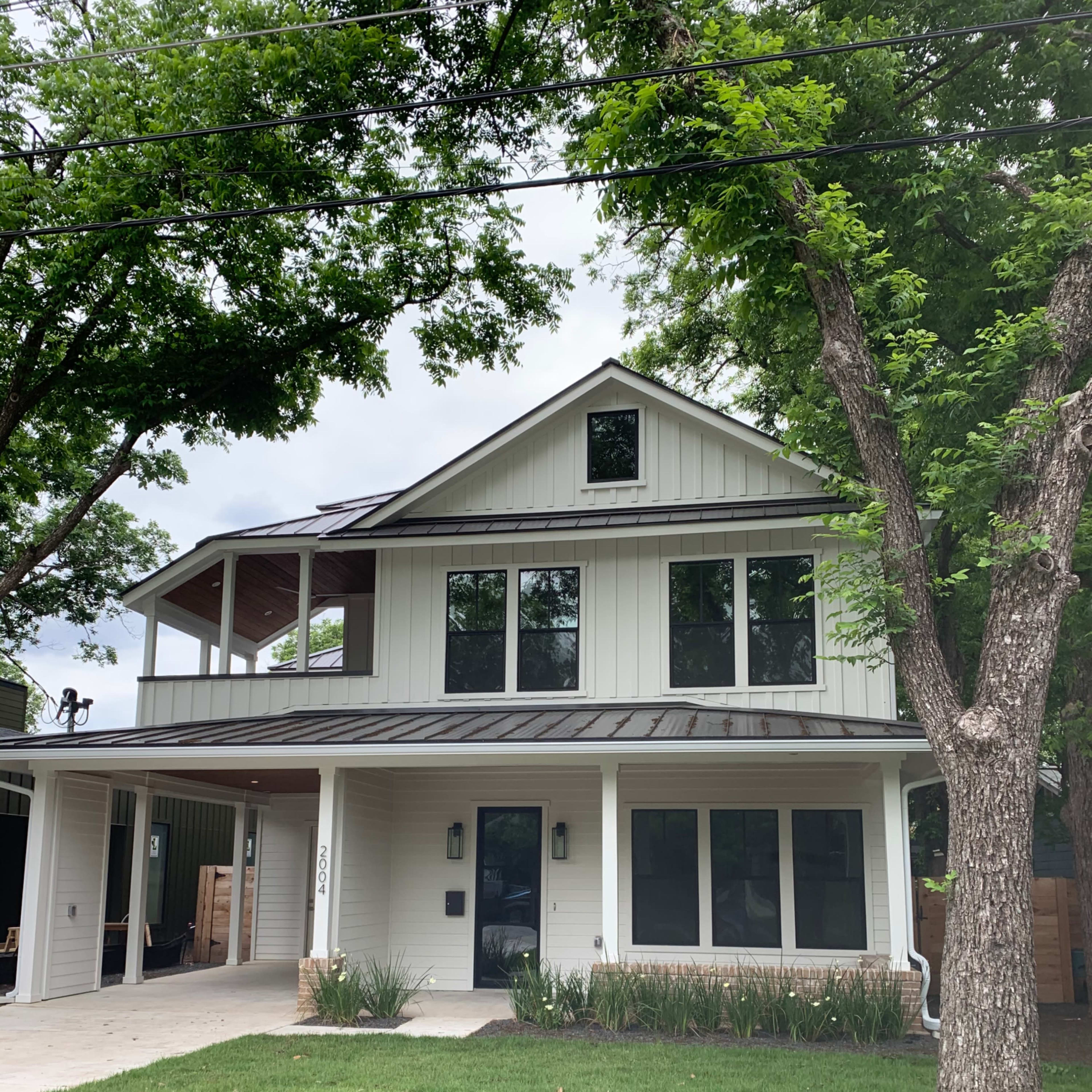 A modern two-story house with a metal roof, large windows, and a front porch, surrounded by greenery and trees.