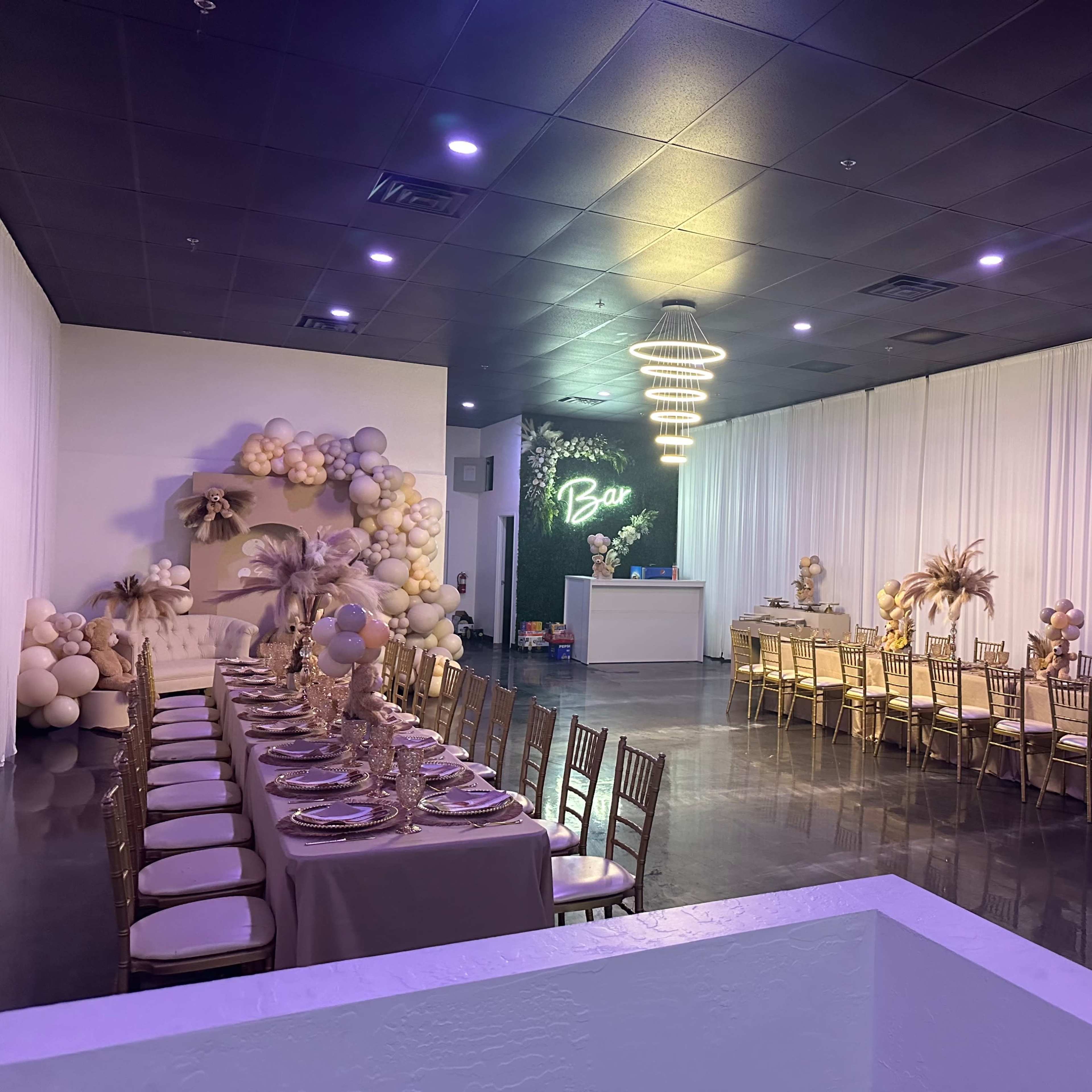 A banquet hall decorated with beige and gold accents, featuring a long table set with dinnerware and a bar area illuminated by pendant lights.