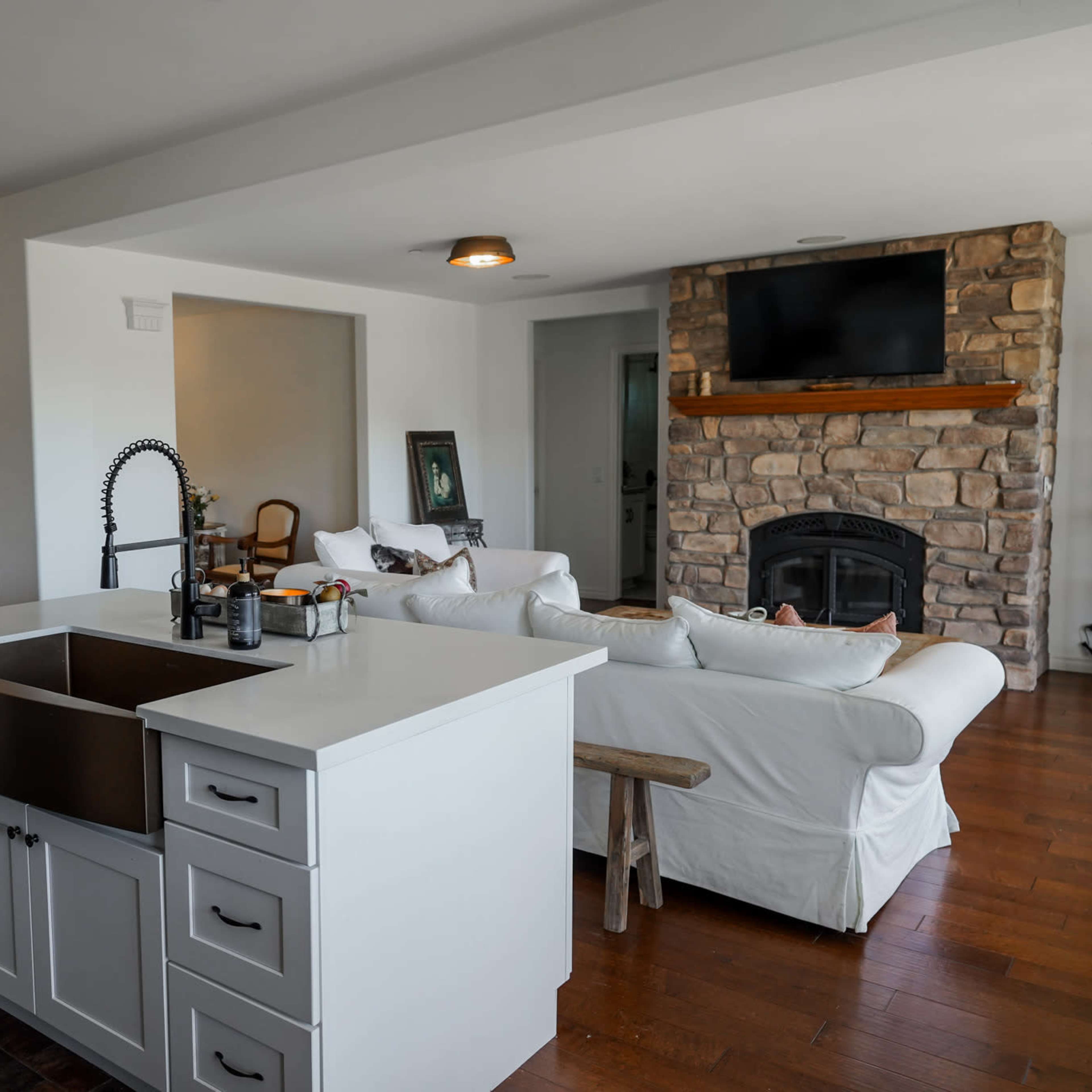 A modern kitchen with a farmhouse sink facing an open living area that features a stone fireplace and a television mounted above it.