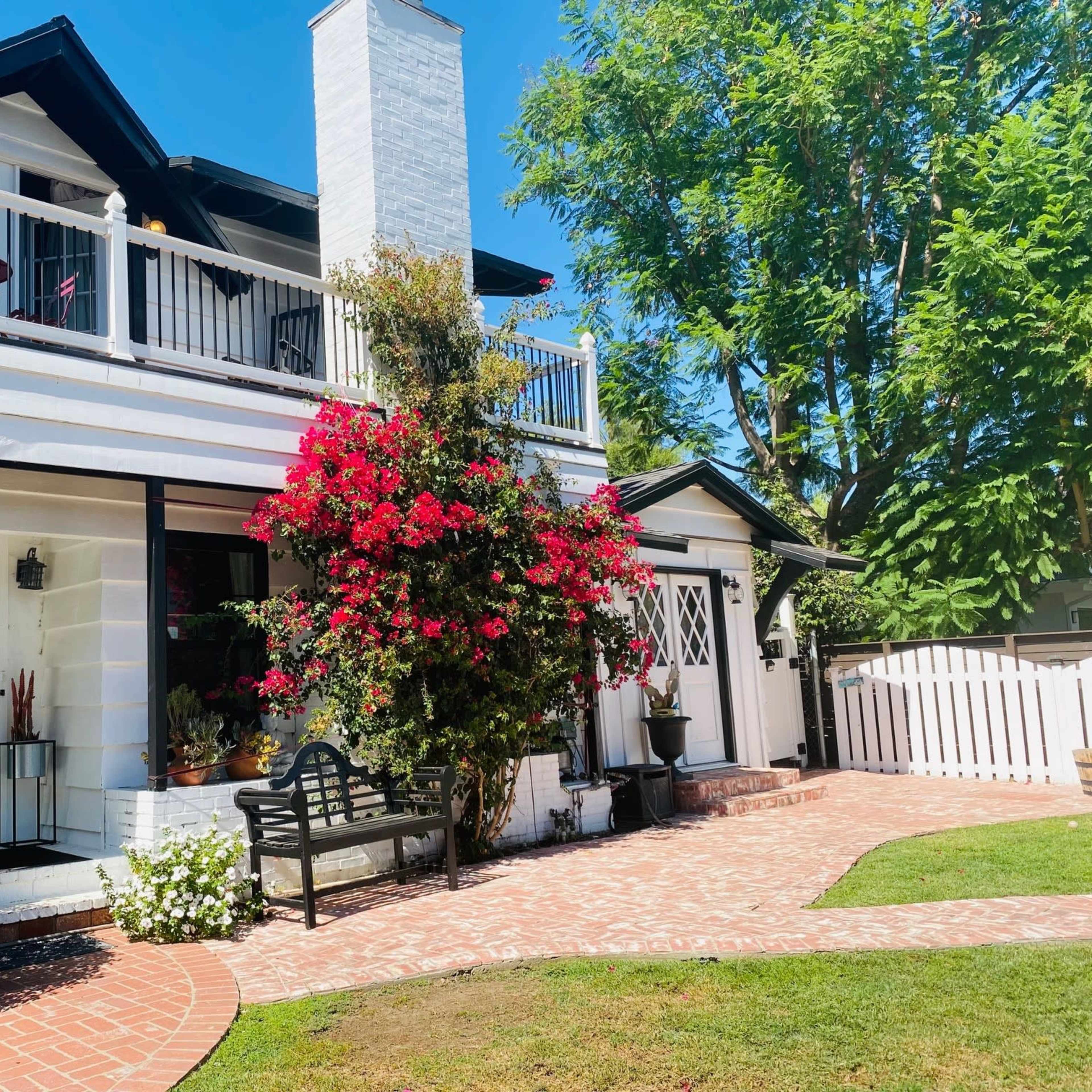 A two-story house with a white exterior, a brick pathway, and a front garden featuring blooming flowers and a bench.