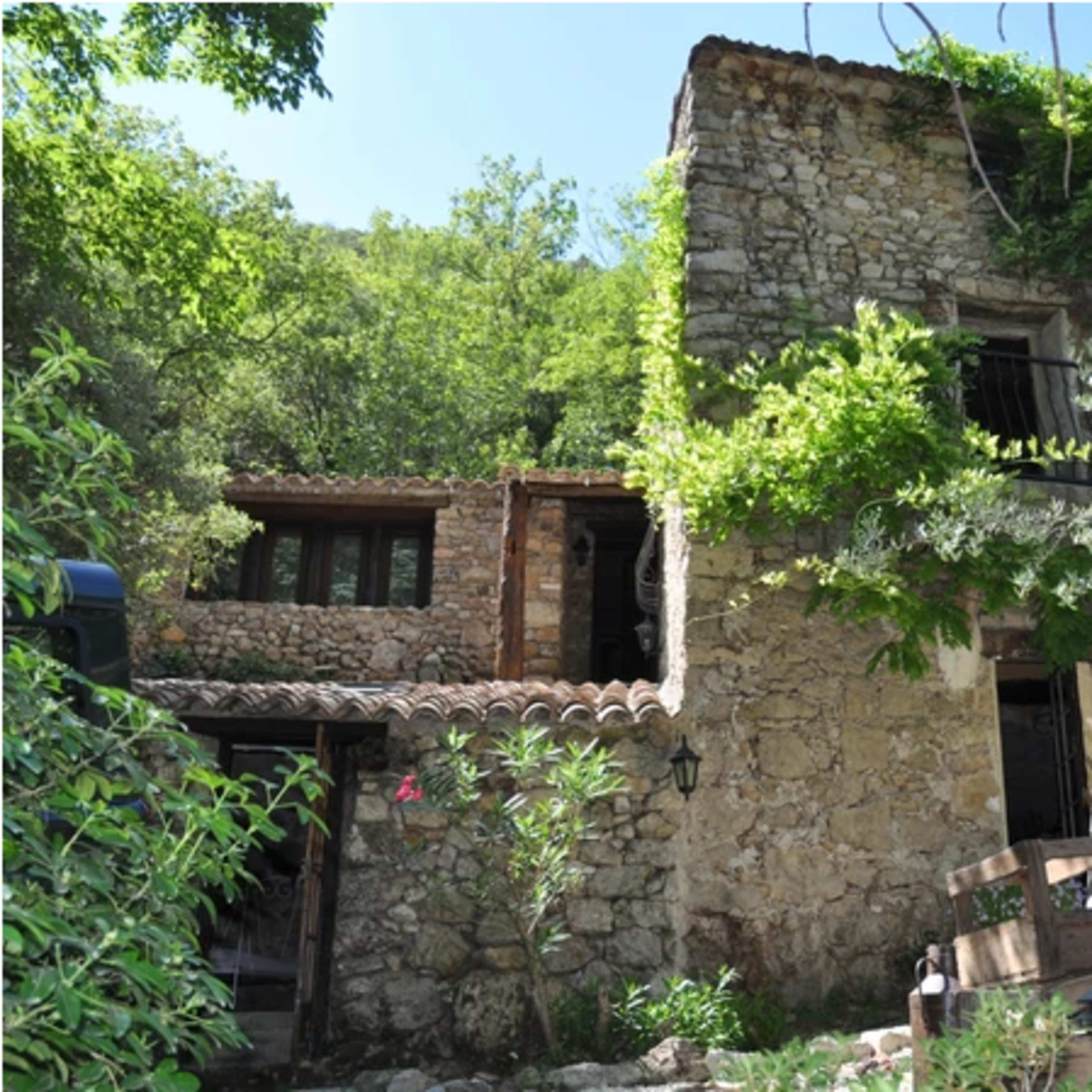 A rustic stone house is surrounded by greenery, with vines climbing the walls and wooden barrels positioned in the foreground.