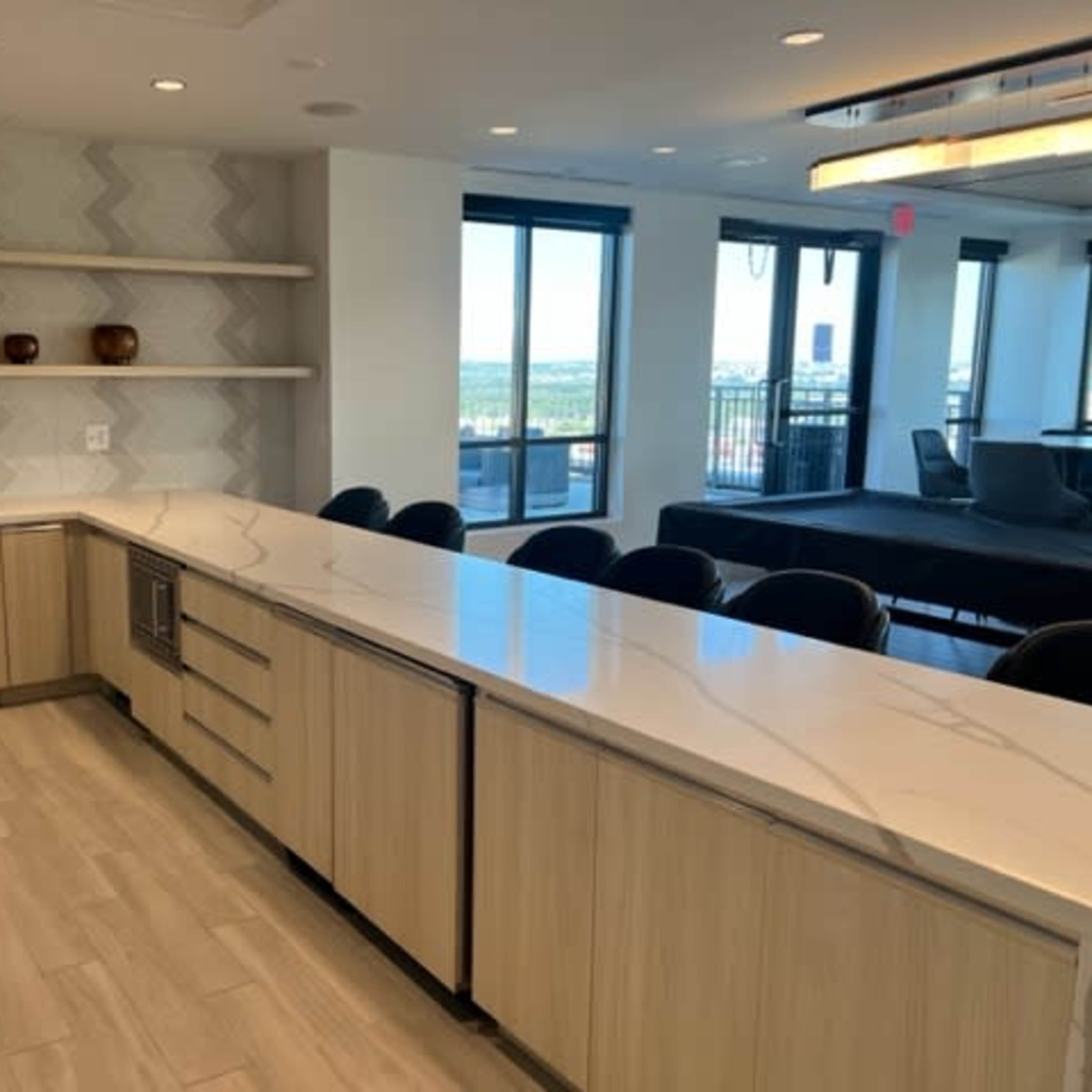 A modern kitchen space with a long countertop, light wood cabinetry, and large windows overlooking a view.