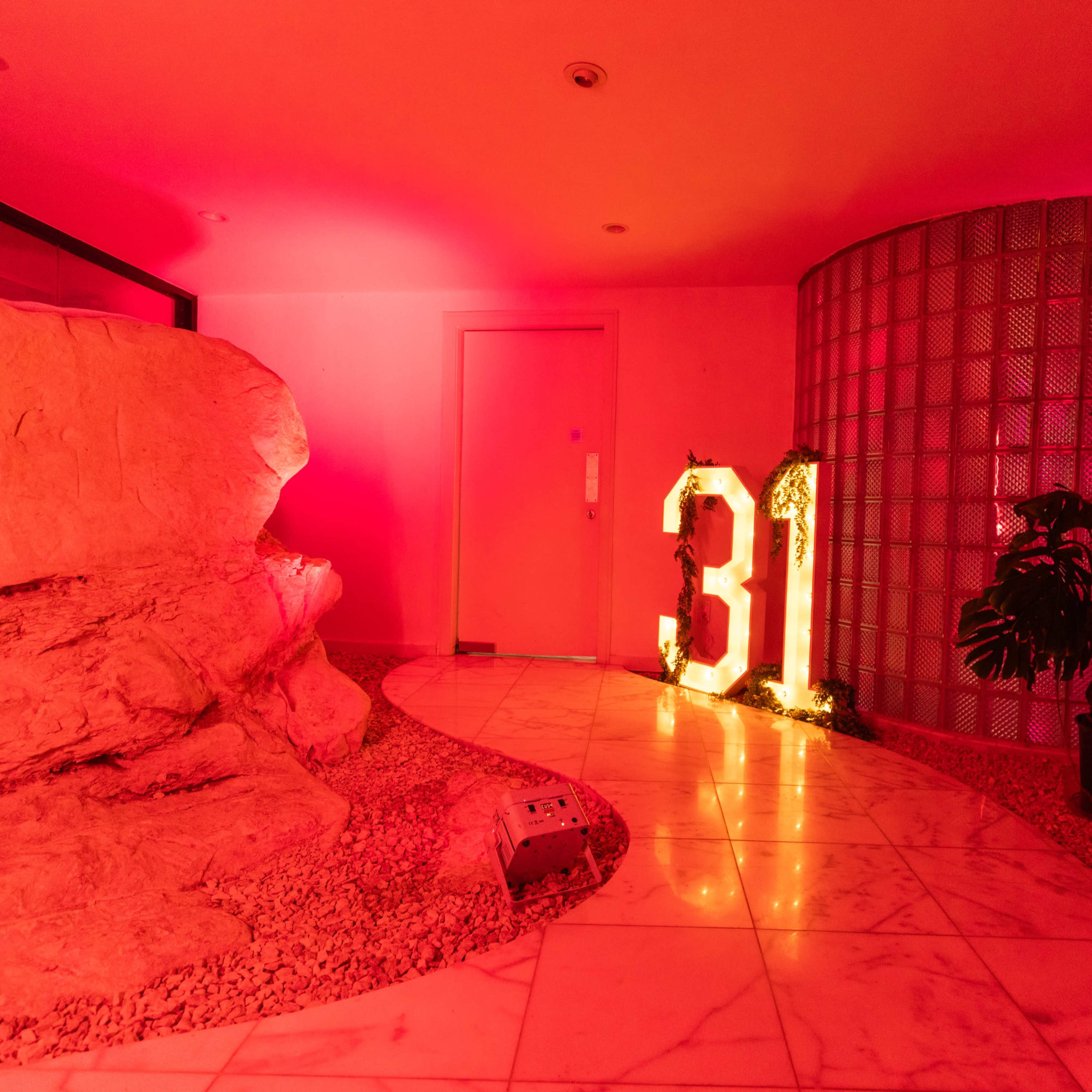 A brightly lit hallway features a large illuminated number 31, with a rocky formation and greenery on either side against a backdrop of red lighting.