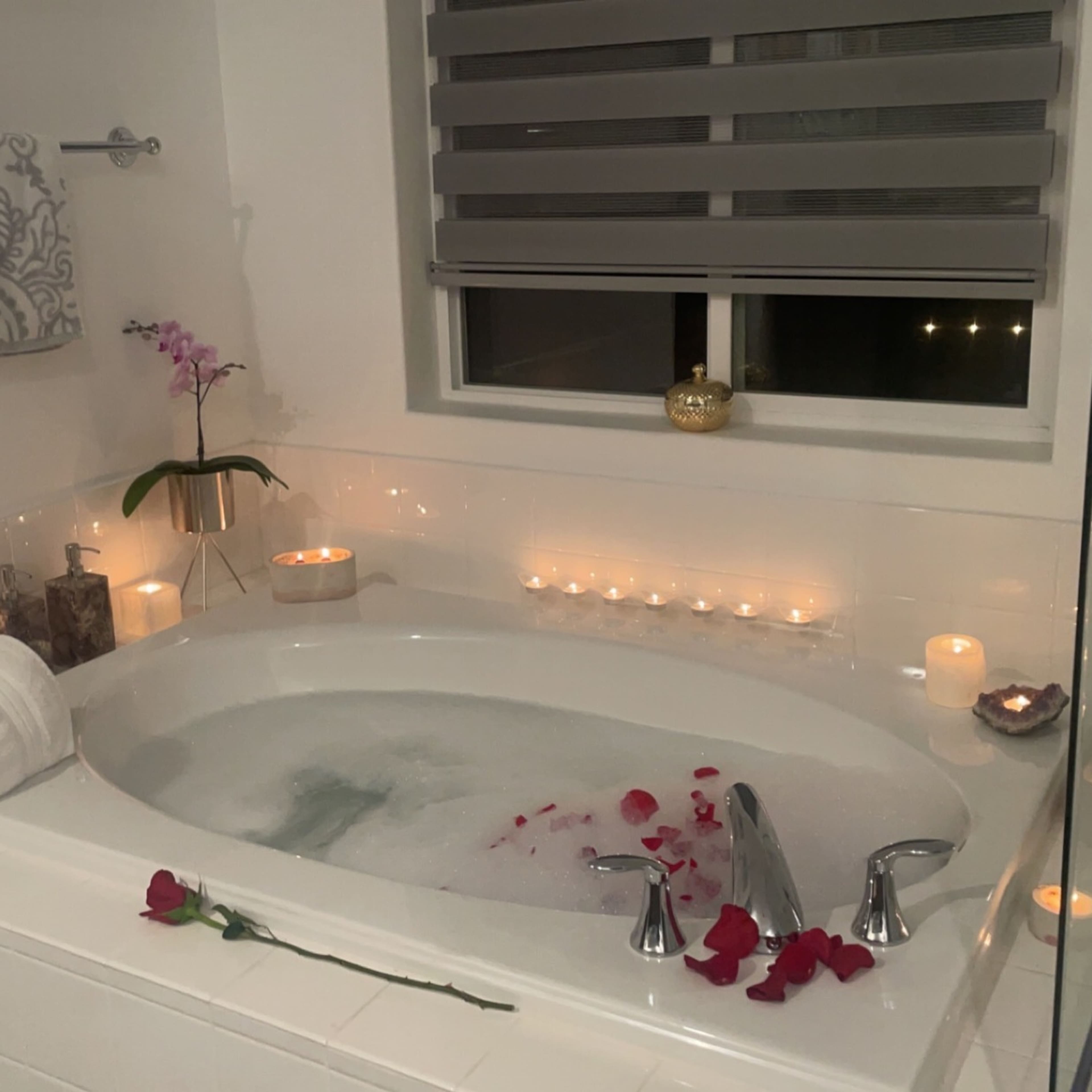 A bathtub surrounded by candles and rose petals is located in a bathroom with a window featuring modern blinds.