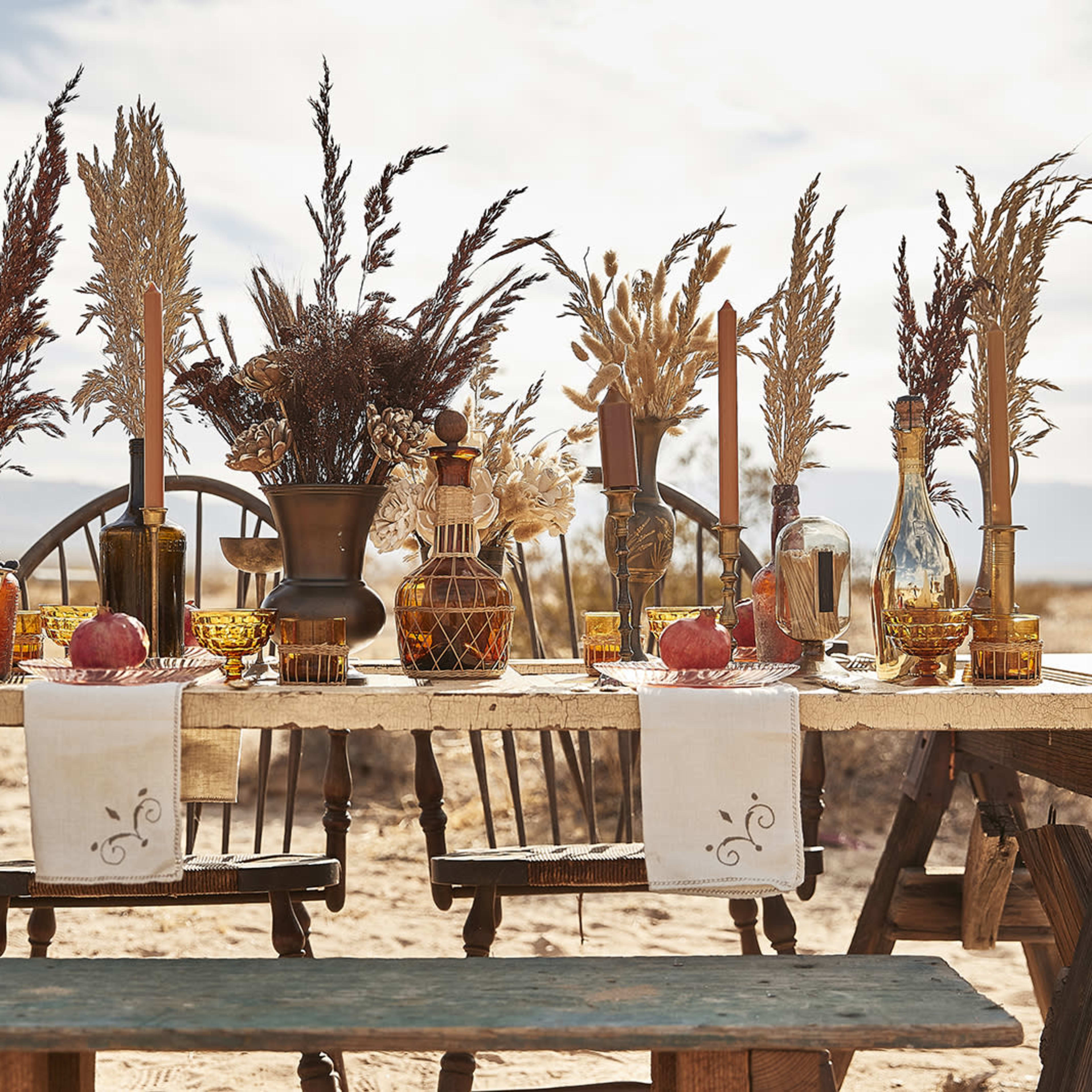 A long wooden table is set outdoors with various decorative items, including glass bottles, candles, and dried floral arrangements in amber tones.