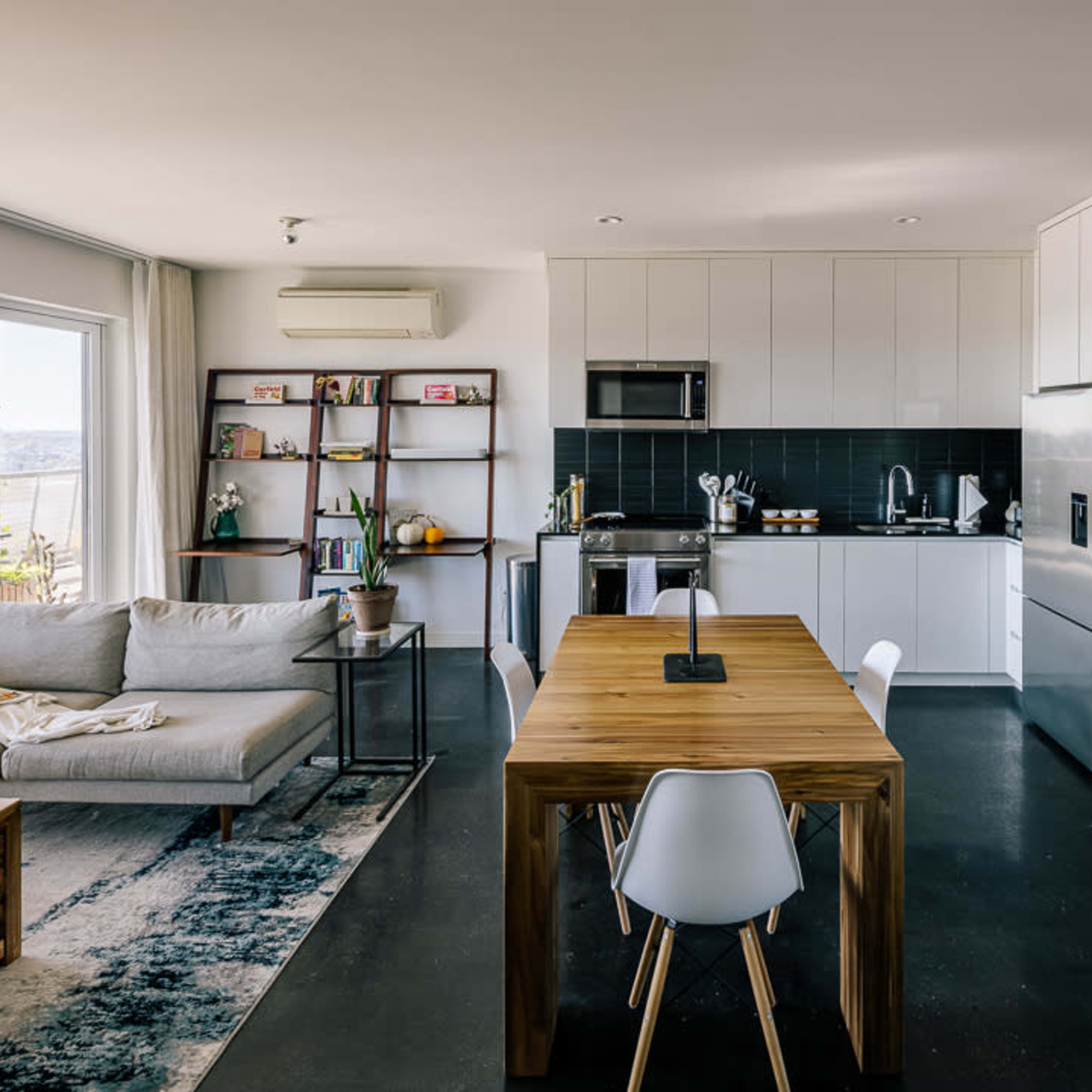 A modern open-concept kitchen and living room featuring a wooden dining table, a light-colored sofa, and large windows with natural light.