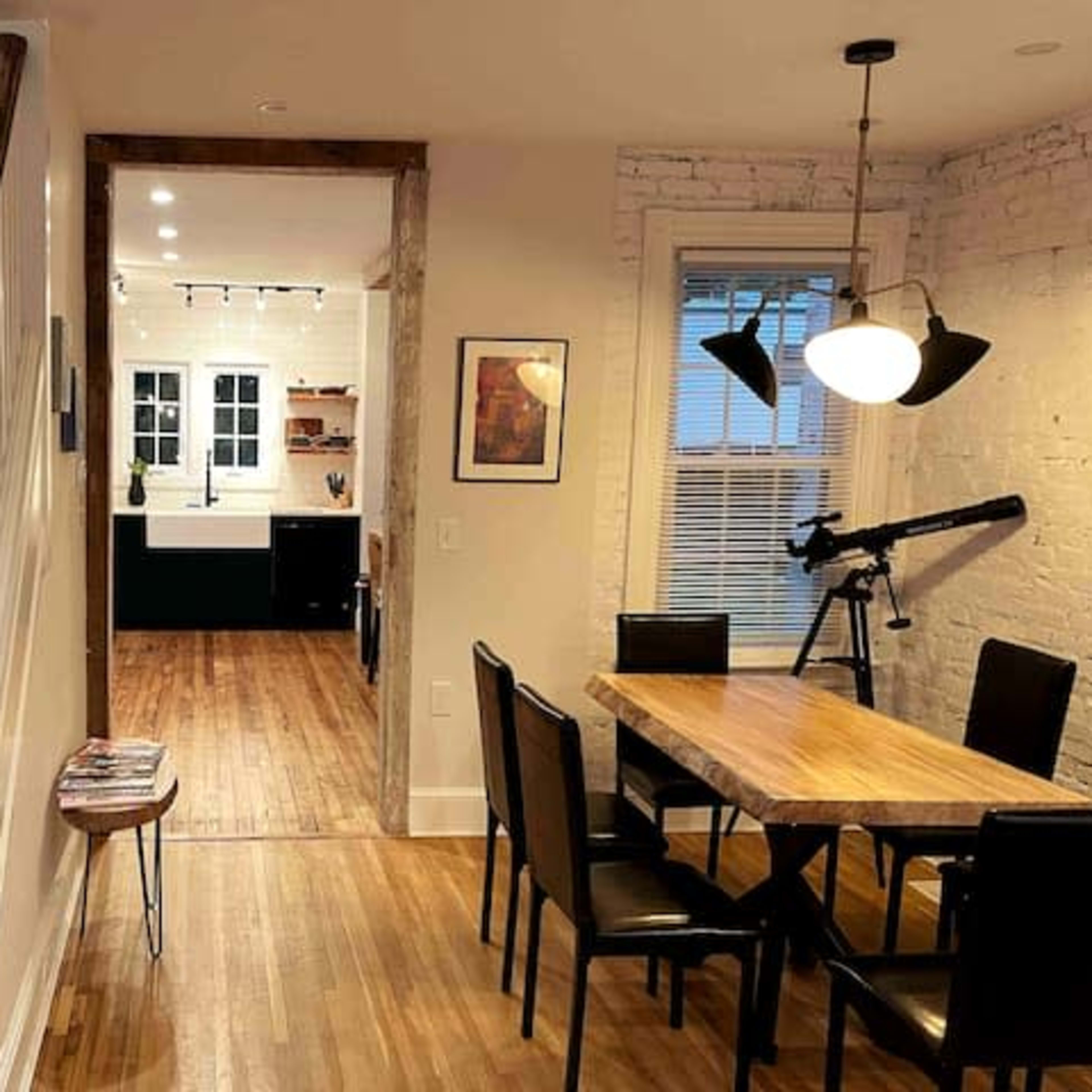 A staircase ascends from a dining area that features a wooden table and black chairs, leading to a kitchen visible through an open doorway.