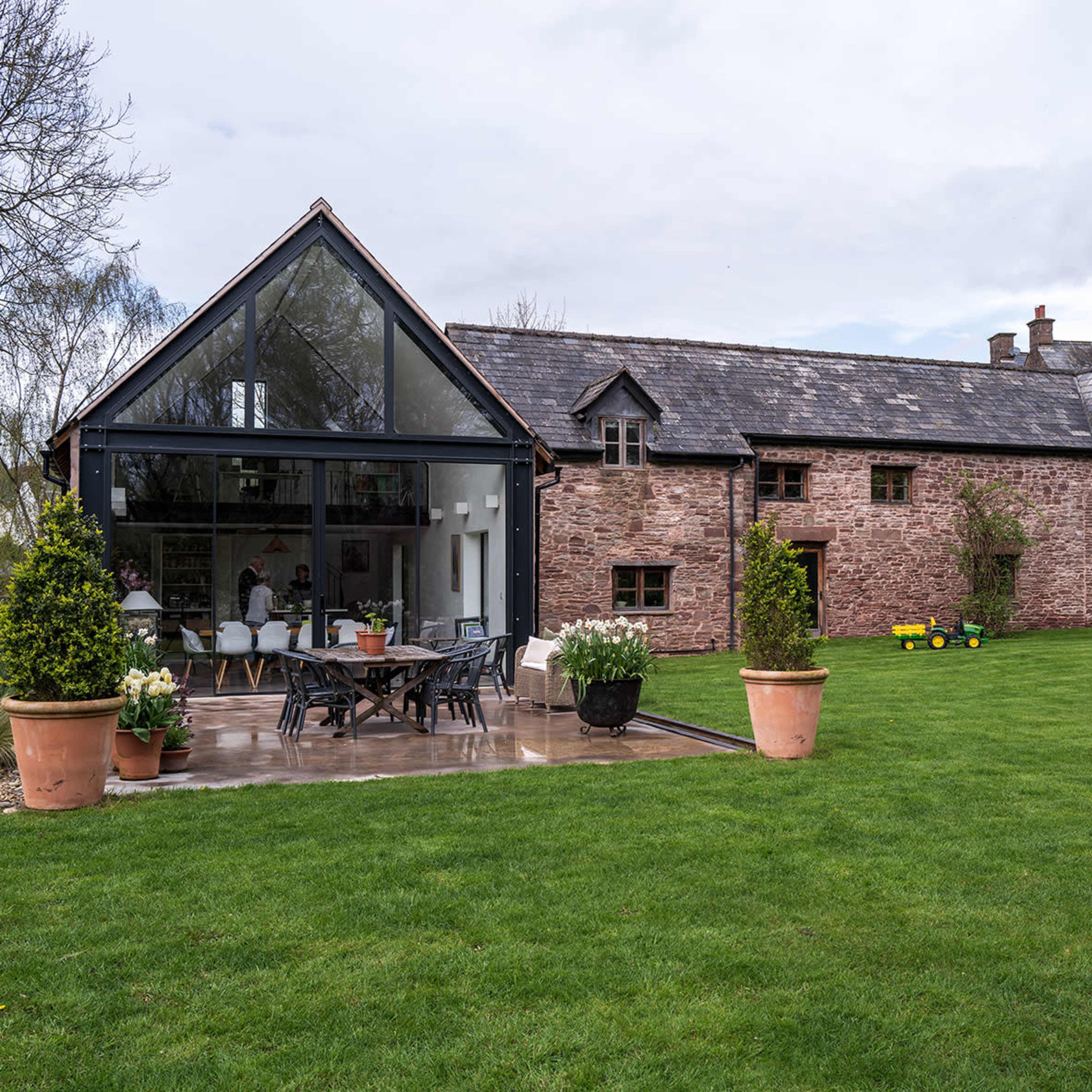 A modern glass extension adjoins a rustic stone farmhouse surrounded by a well-maintained lawn and garden.