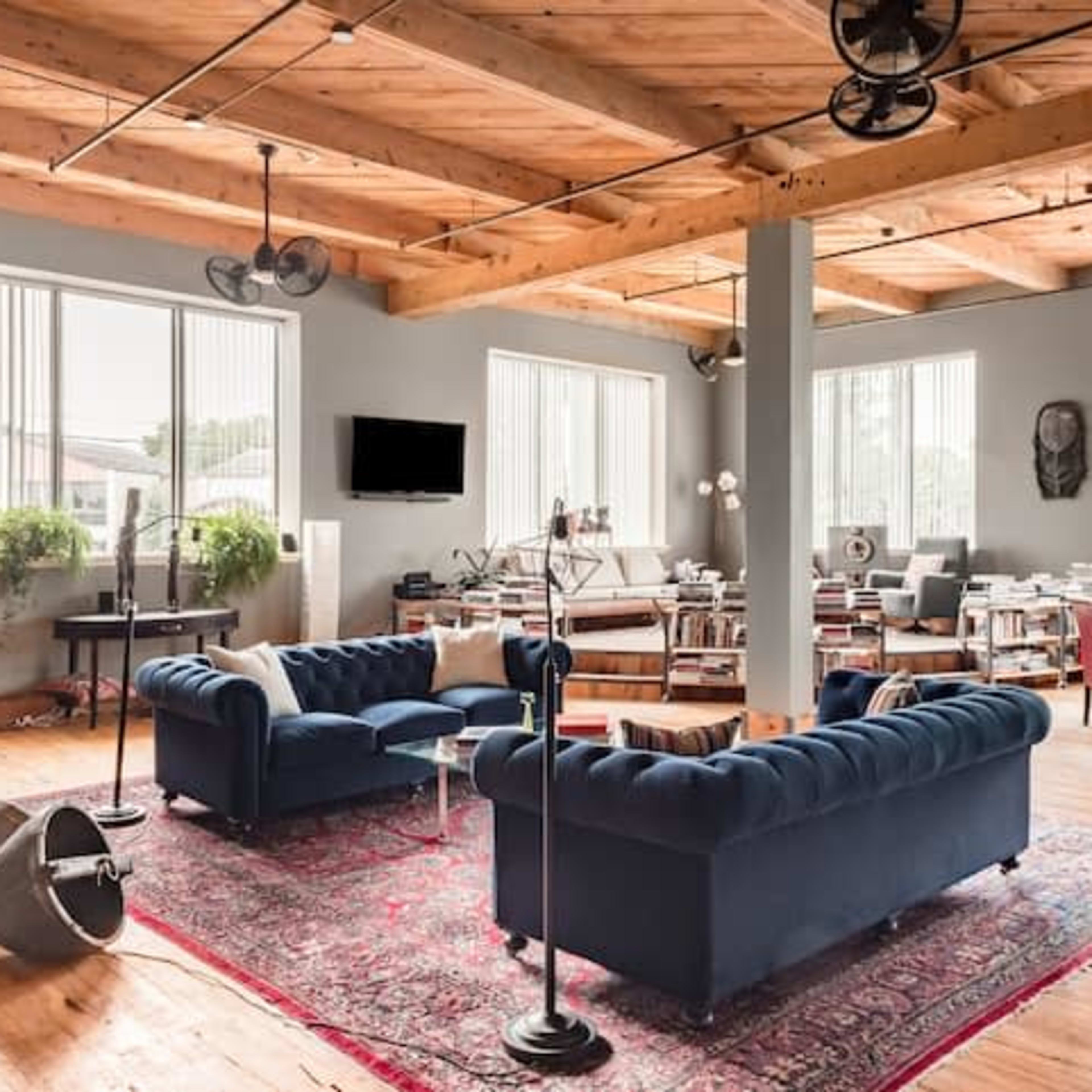 A spacious, modern open-concept living area with wooden beams, a large area rug, and various seating arrangements near numerous windows.
