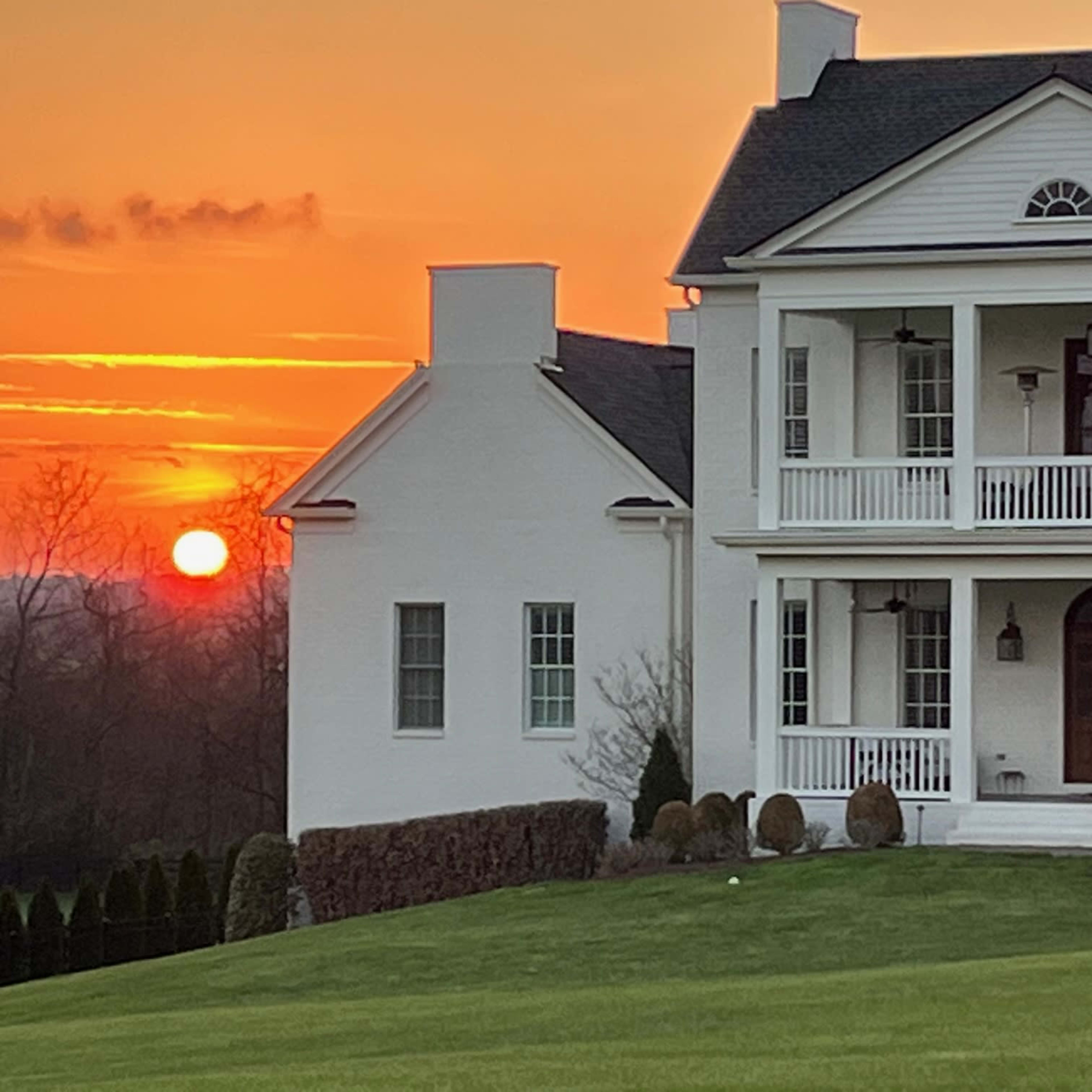 A white house with multiple balconies stands against a backdrop of a sunset, highlighting the sun partially obscured by the horizon.