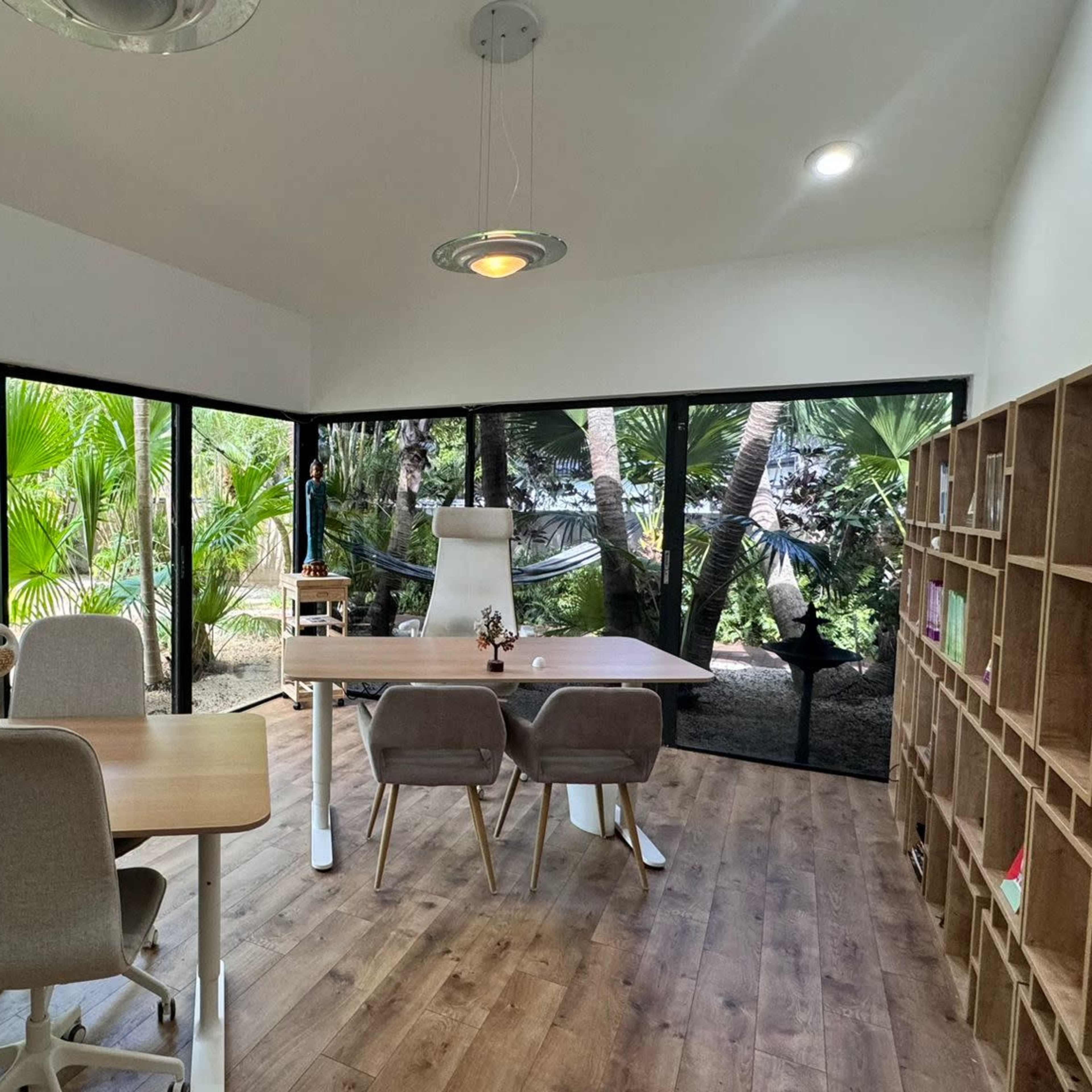 A bright, modern office space with large windows overlooking a tropical garden, featuring a table, chairs, and wooden shelving.
