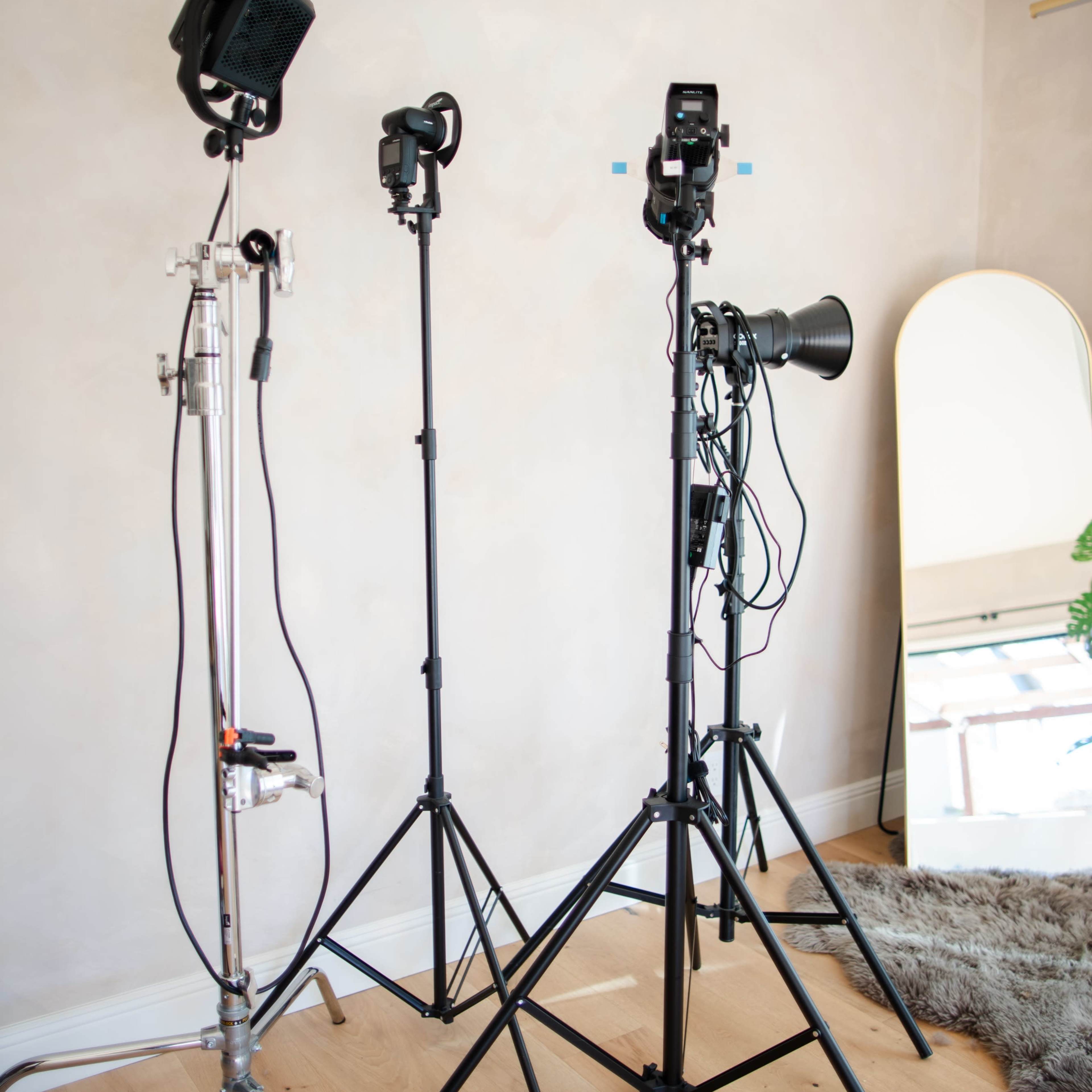 Three photography light stands positioned near a wall with a mirror and wooden flooring.