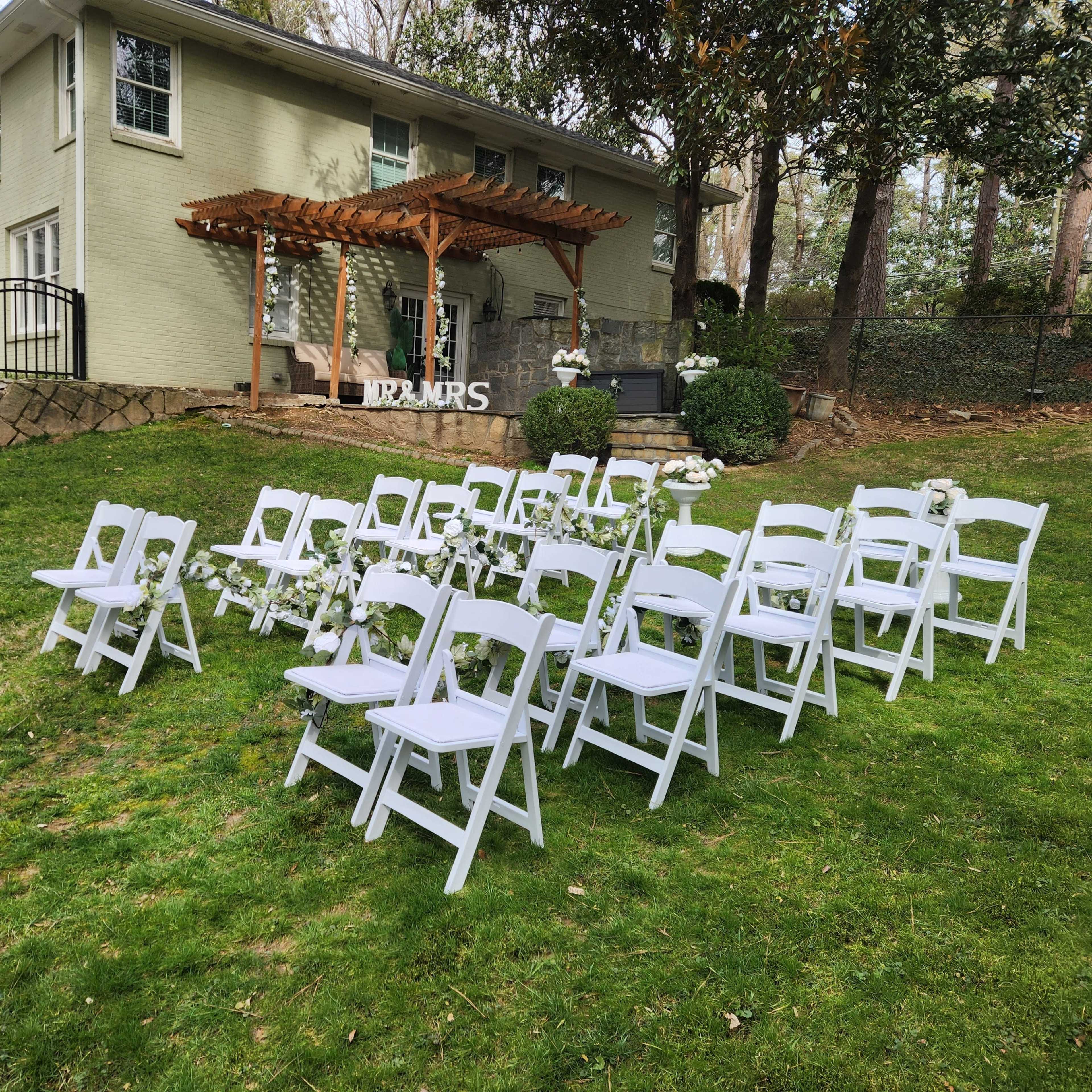 A wedding setup featuring rows of white folding chairs arranged on a green lawn in front of a house with a wooden pergola.
