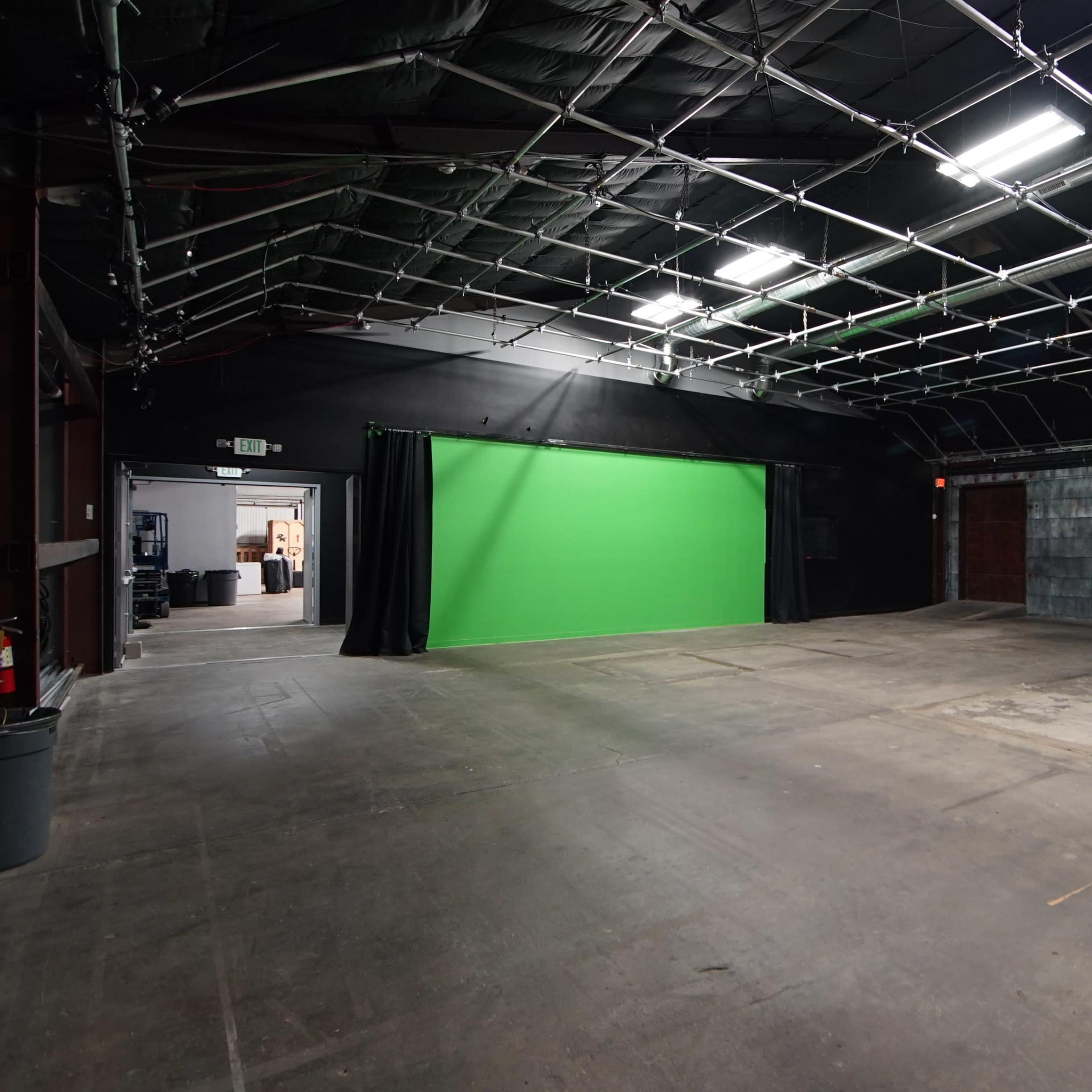 An interior of a large studio space with a green screen at one end and exposed metal walls.