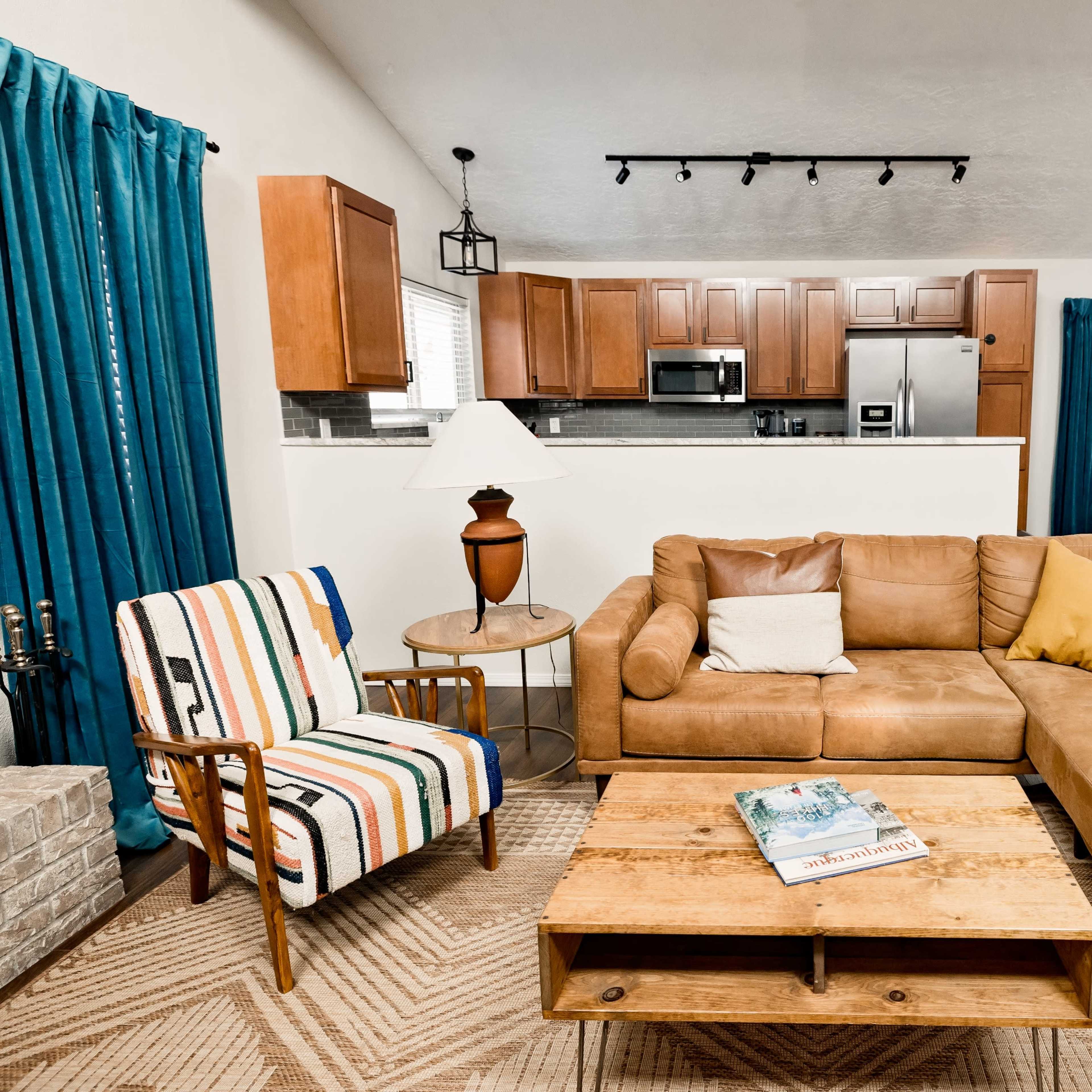 A cozy living room features a brown leather couch, a striped armchair, a coffee table with a magazine, and a kitchen area visible in the background.